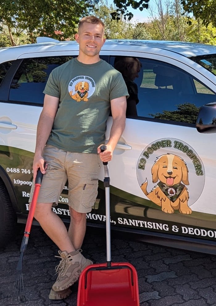 A man in uniform standing in front of a branded service vehicle, holding a rake and dustpan suitable for Dog Waste Removal (pooper scooping). The vehicle has graphics of the K9 Pooper Troopers logo and company information.