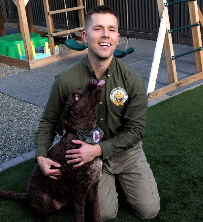 A smiling man in uniform from K9 Pooper Troopers kneeling on grass with a brown, curly-coated dog attempting to lick him in an outdoor play area with swings and play structures.