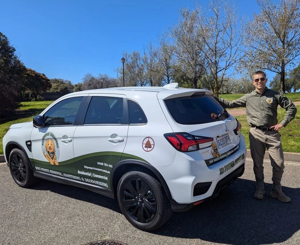 A smiling man in a uniform standing next to a white K9 Pooper Troopers dog waste removal vehicle parked on a paved road in a park with green grass and trees. The vehicle has graphics of their logo, which features a cute dog, and the business details.