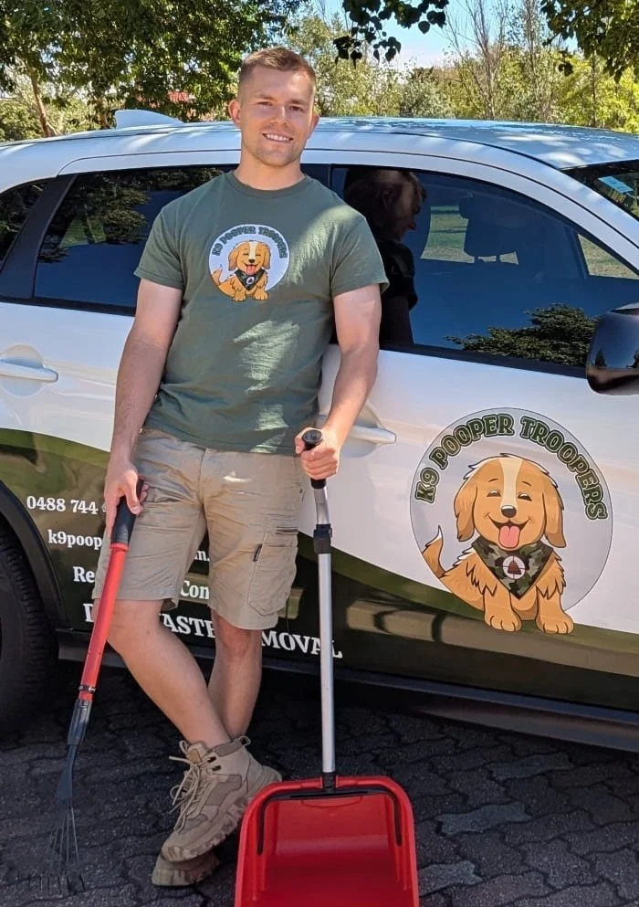 A man in uniform standing in front of a branded service vehicle, holding a rake and dustpan suitable for Dog Waste Removal (pooper scooping). The vehicle has graphics of the K9 Pooper Troopers logo and company information.