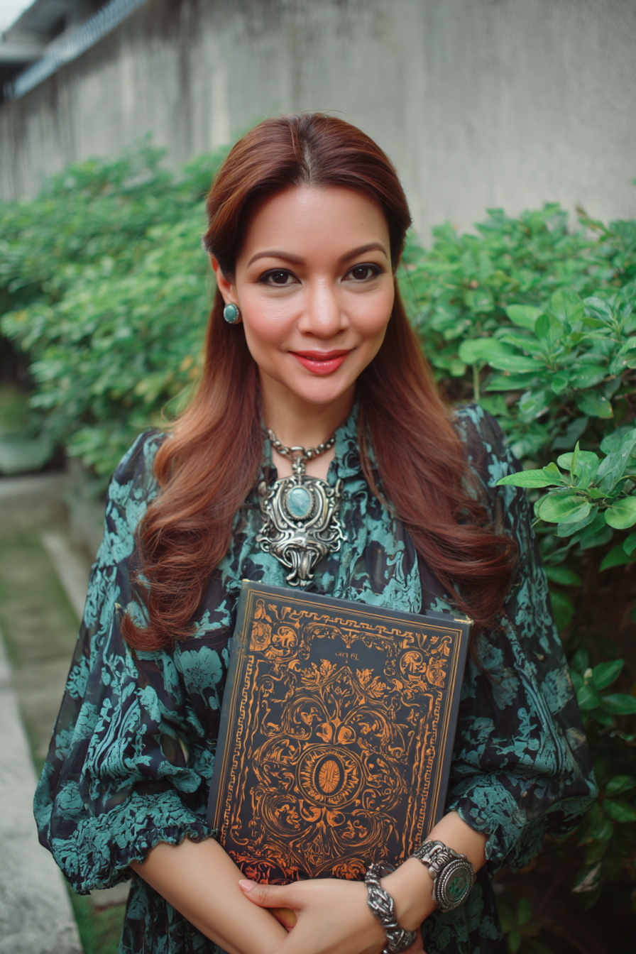 A woman with long, wavy brown hair wearing a patterned blue and black dress holds an ornate black and gold book. She has turquoise earrings, a large statement necklace, a silver bracelet, and a large silver watch. She is standing outdoors near green bushes and a concrete wall, smiling at the camera.