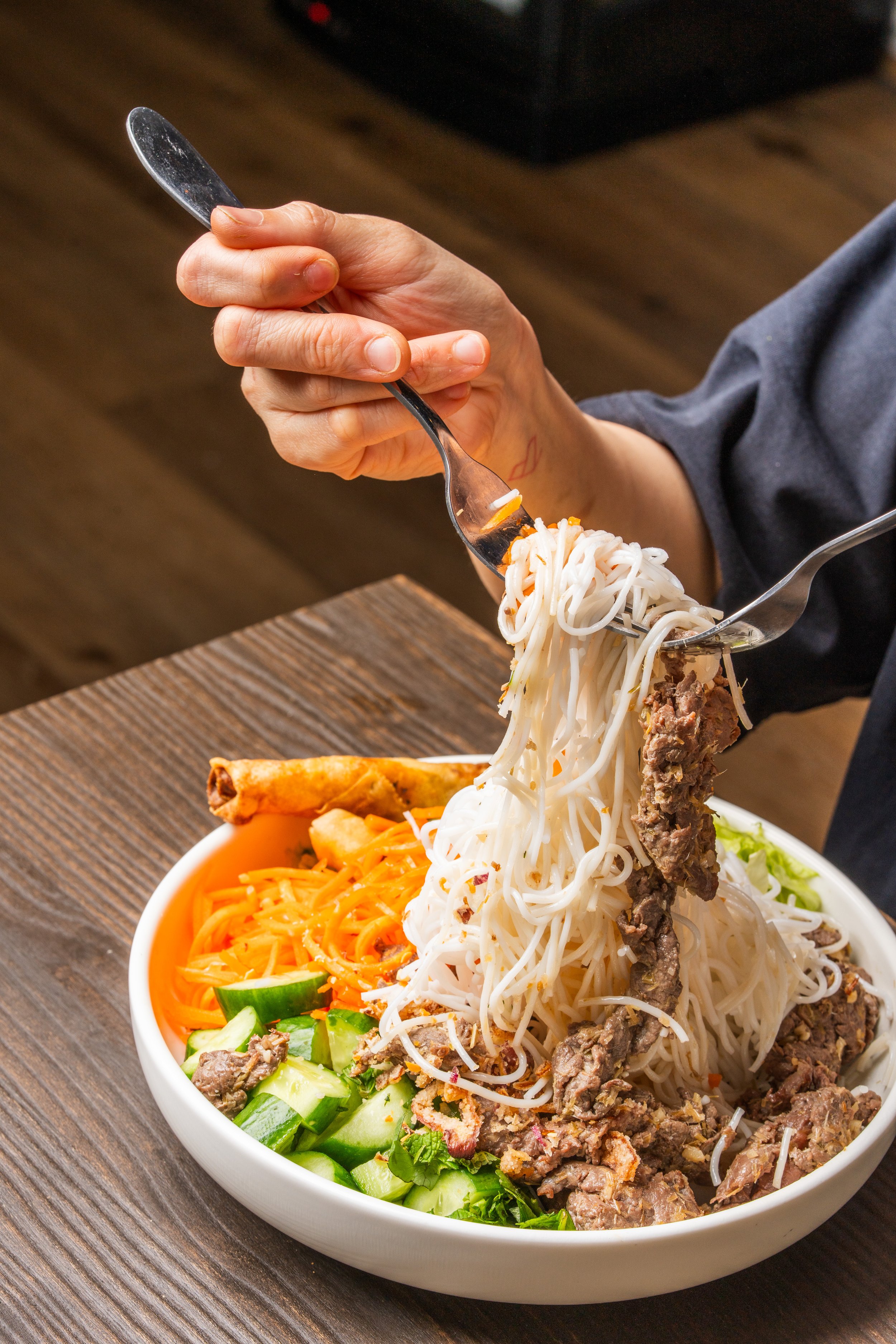 A person enjoying a large serve of vermicelli noodles, meat, fresh vegetables, accompanied by a crispy spring roll