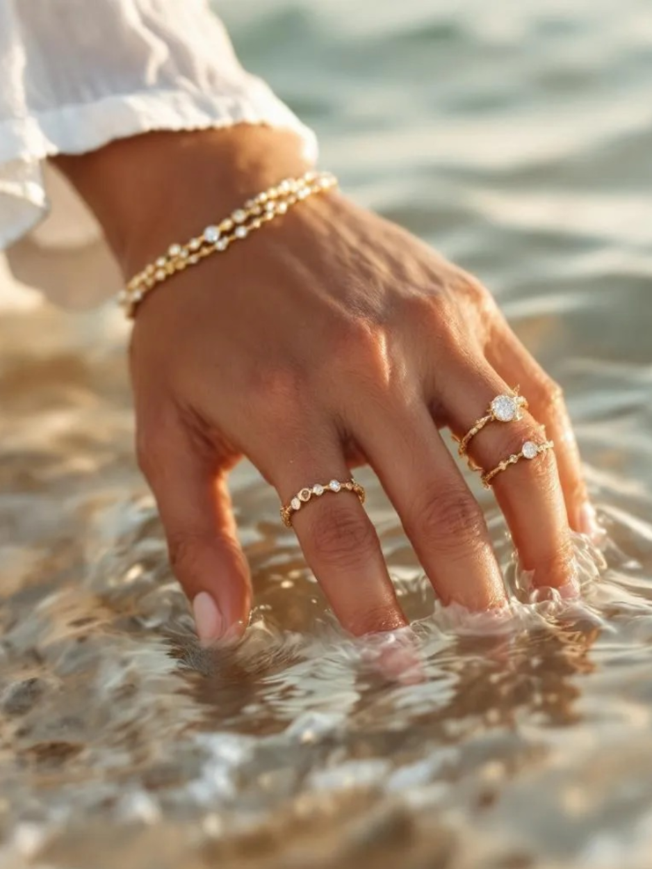 A hand adorned with gold rings and bracelets dipped in clear ocean water, capturing the quiet luxury and water-friendly elegance of Krisalini jewelry.