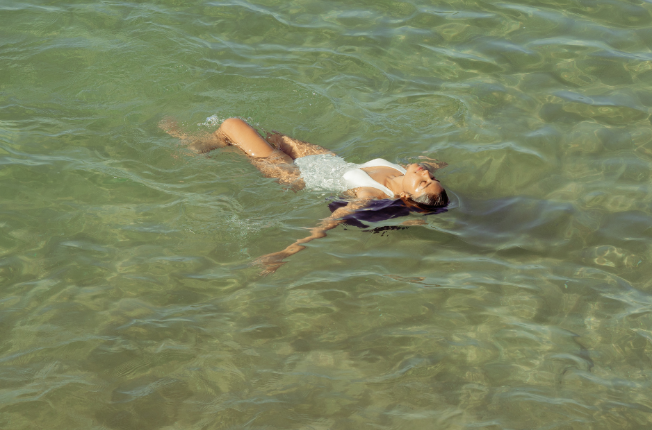 Woman floating peacefully in clear coastal water wearing minimal jewelry, embodying slow living and quiet luxury.