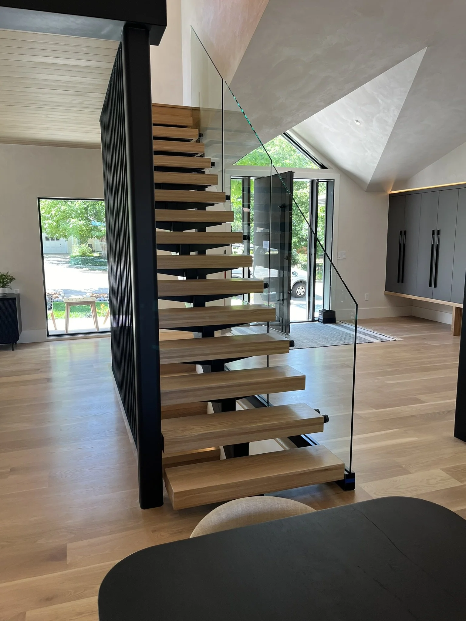 Interior of a modern home featuring a floating wooden staircase with black metal supports, large windows bringing in natural light, and a gray wall with built-in storage.