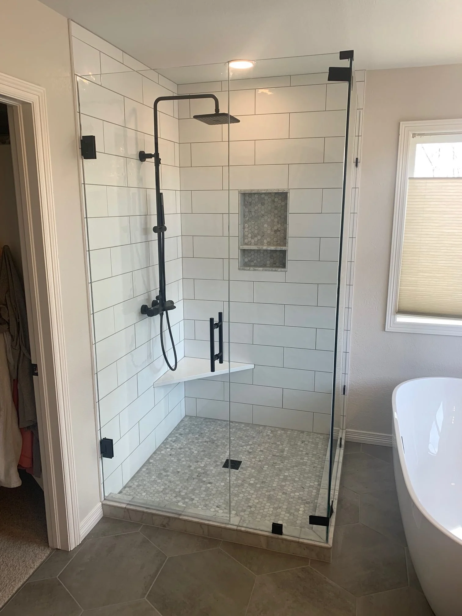 A modern walk-in shower with white large rectangular tiles, a black showerhead and fixtures, built-in niche with smaller gray hexagon tiles, and a glass door, in a bathroom with gray floor tiles and a window with blinds.