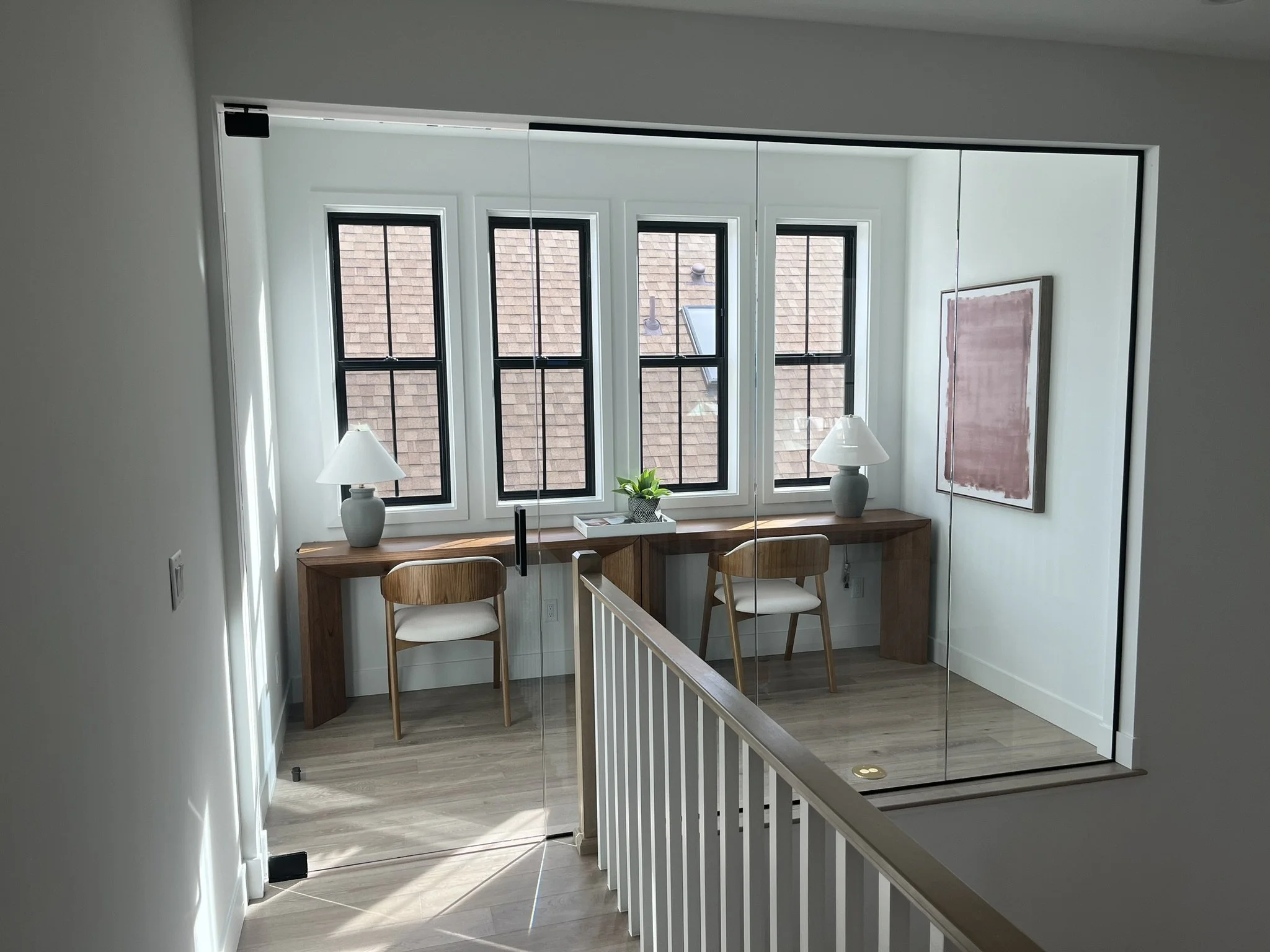 Interior of a room with large windows, two white lamps, a wooden table, and two wooden chairs. There is a plant and a pink artwork on the wall. The photo is reflected in a mirror.