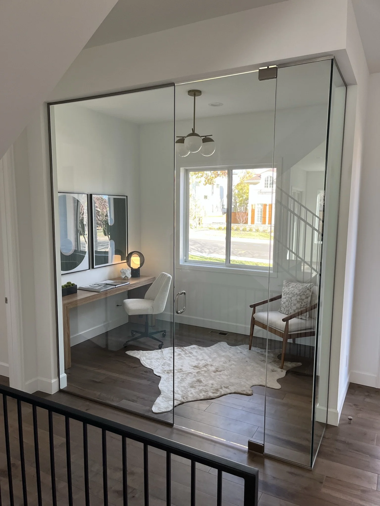 A room viewed through glass doors with a desk, a mirror, a chair, a rug, and a window showing houses outside.