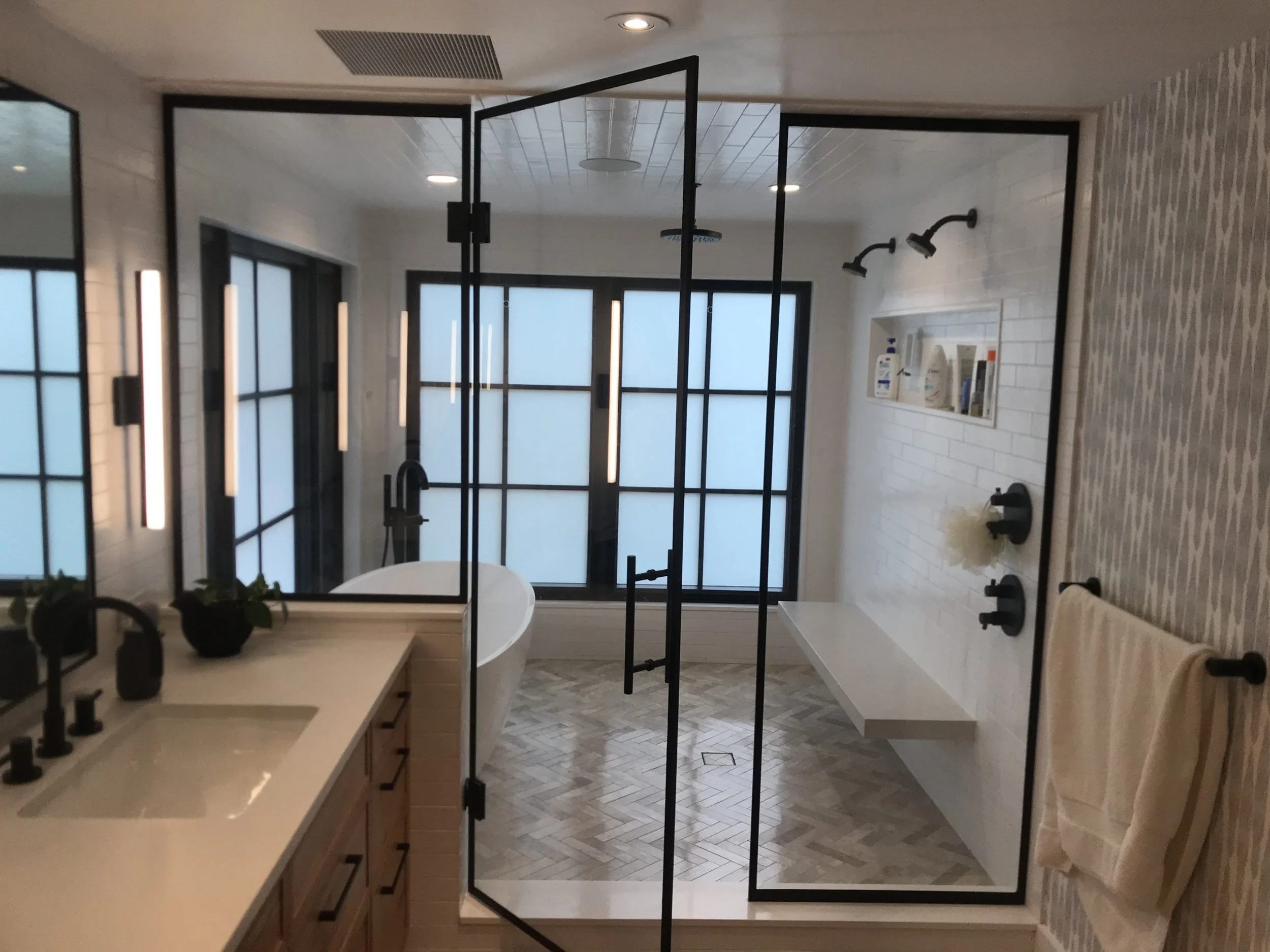 A spacious modern bathroom with a walk-in shower enclosed by black metal frames and frosted glass panels, a white bathtub, a vanity with a vessel sink, a large mirror, and modern fixtures, with natural light coming through large frosted windows.