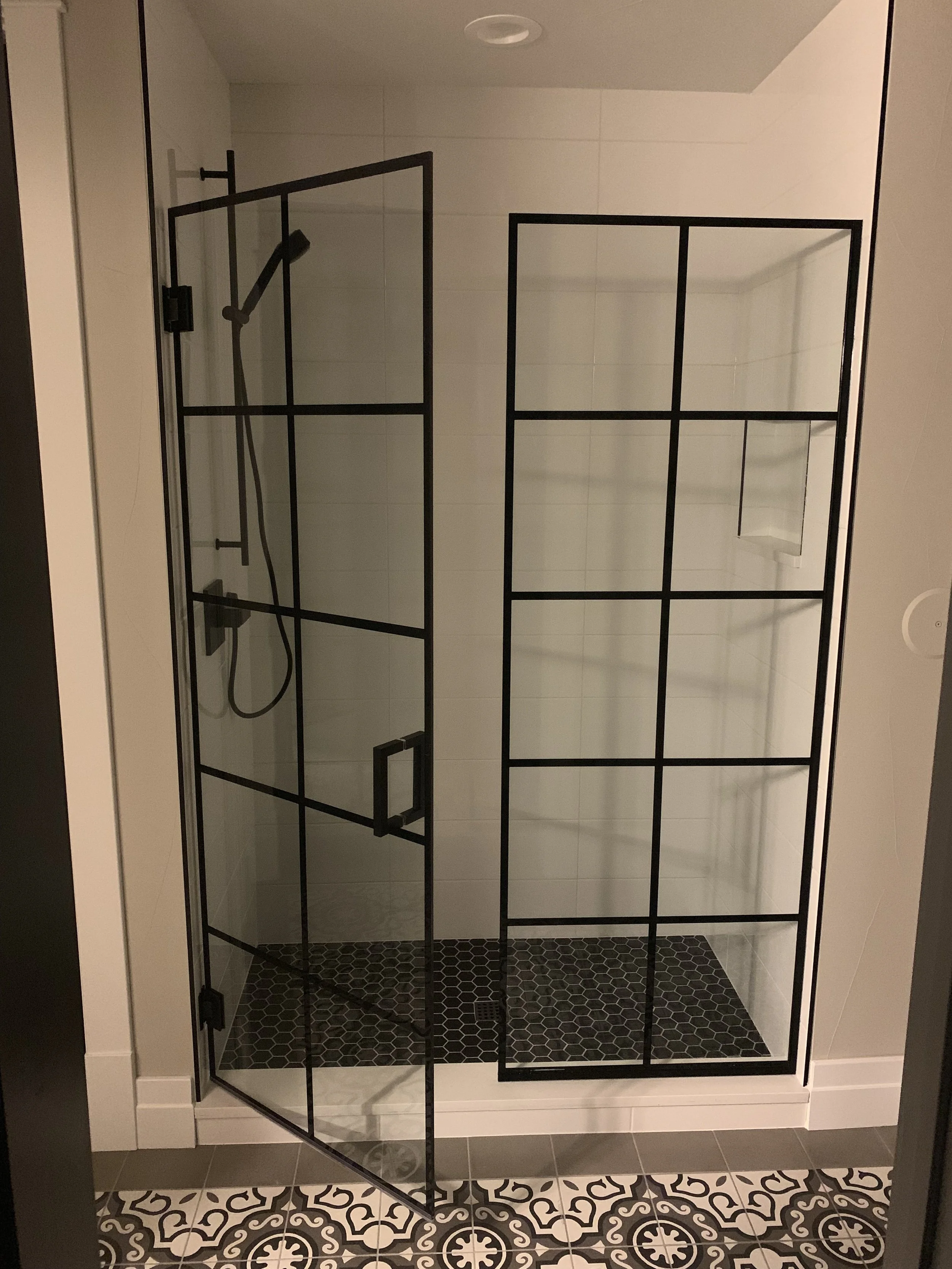 Modern bathroom shower with clear glass walls and black metal frame, white tiled walls, black patterned floor tiles, and a rainfall showerhead.