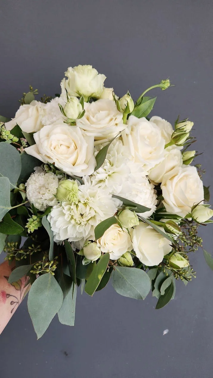 A wedding bouquet of white roses, carnations, and other white flowers with green leaves and eucalyptus, held in a person's hand with a tattoo on their wrist against a gray background.