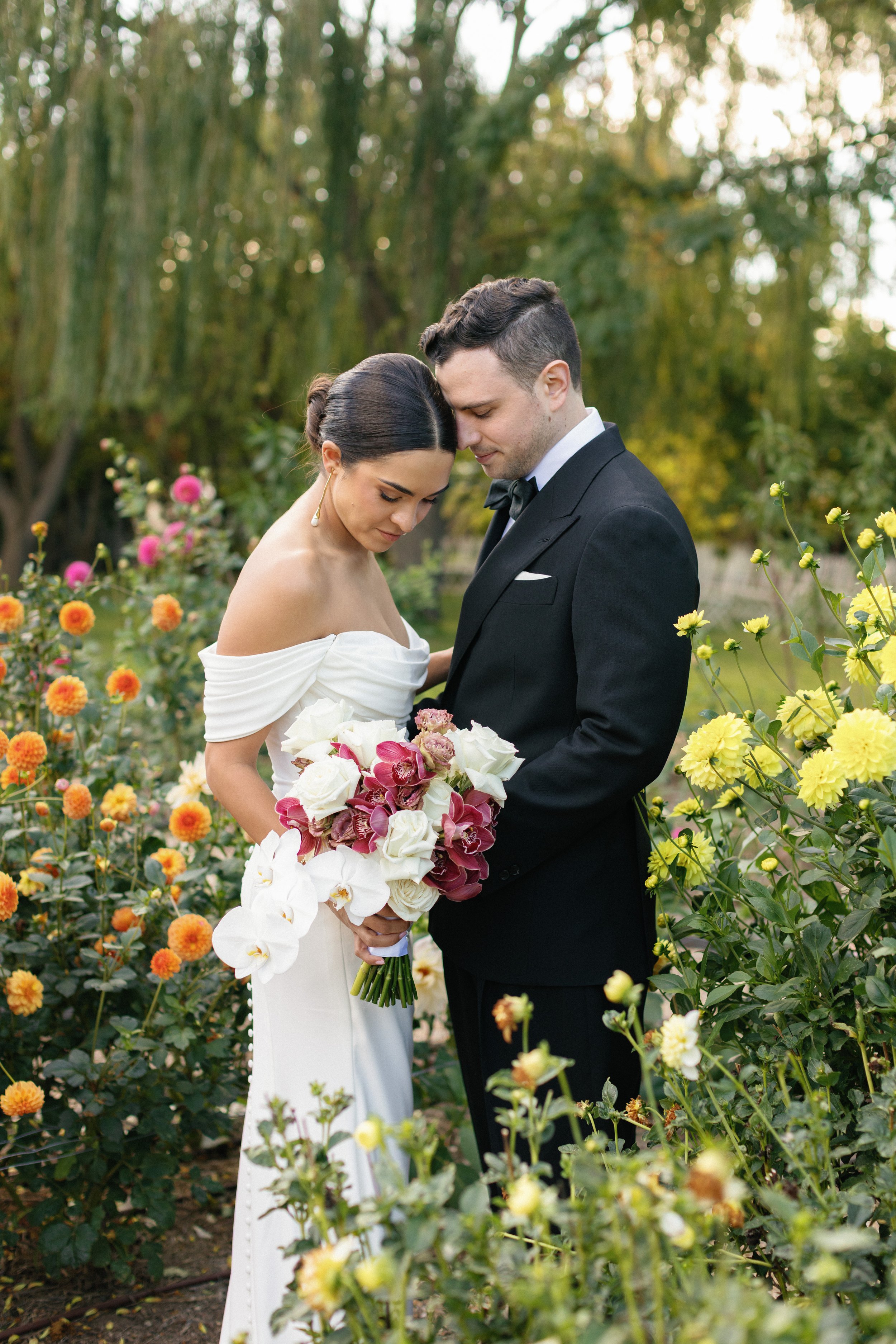 A bride and groom embrace each other in a wedding photo during sunset, with the bride holding a bouquet of flowers and wearing a veil, in an outdoor setting with trees in the background.
