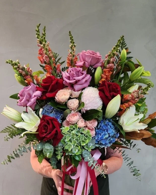 A person holding a large centrepiece of mixed flowers including roses, lilies, hydrangeas, and greenery, tied with a pink and white ribbon.