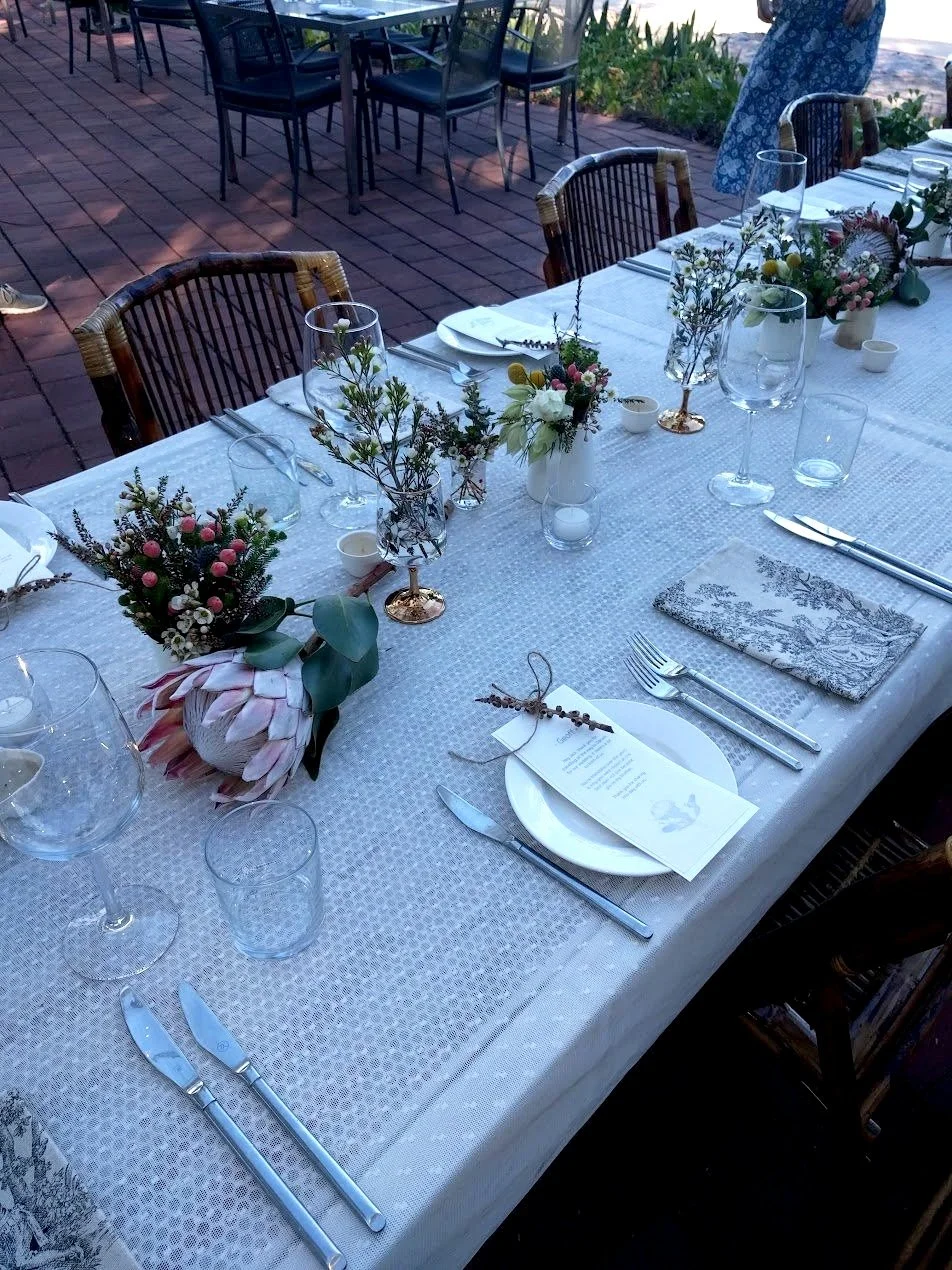 Elegant outdoor dining table set with white tablecloth, floral centerpieces, wine glasses, napkins, and silverware, on a patio with brick flooring.