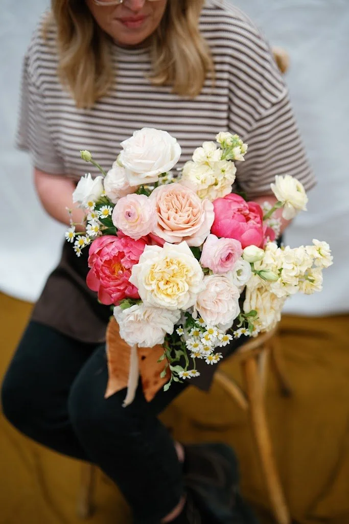 A woman holding a bouquet of various pink, white, and cream flowers, including roses and peonies, while sitting on a wooden chair.