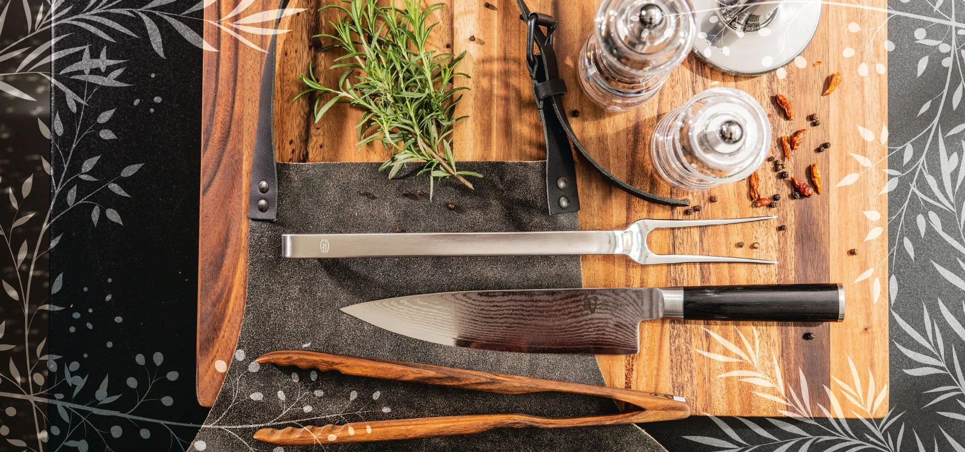 Kochutensilien und Kräuter auf einem Holzbrett, darunter ein Küchenmesser, ein Fleischgabel, Pfeffer- und Salzstreuer, Wacholderbeeren, und frische Kräuter.