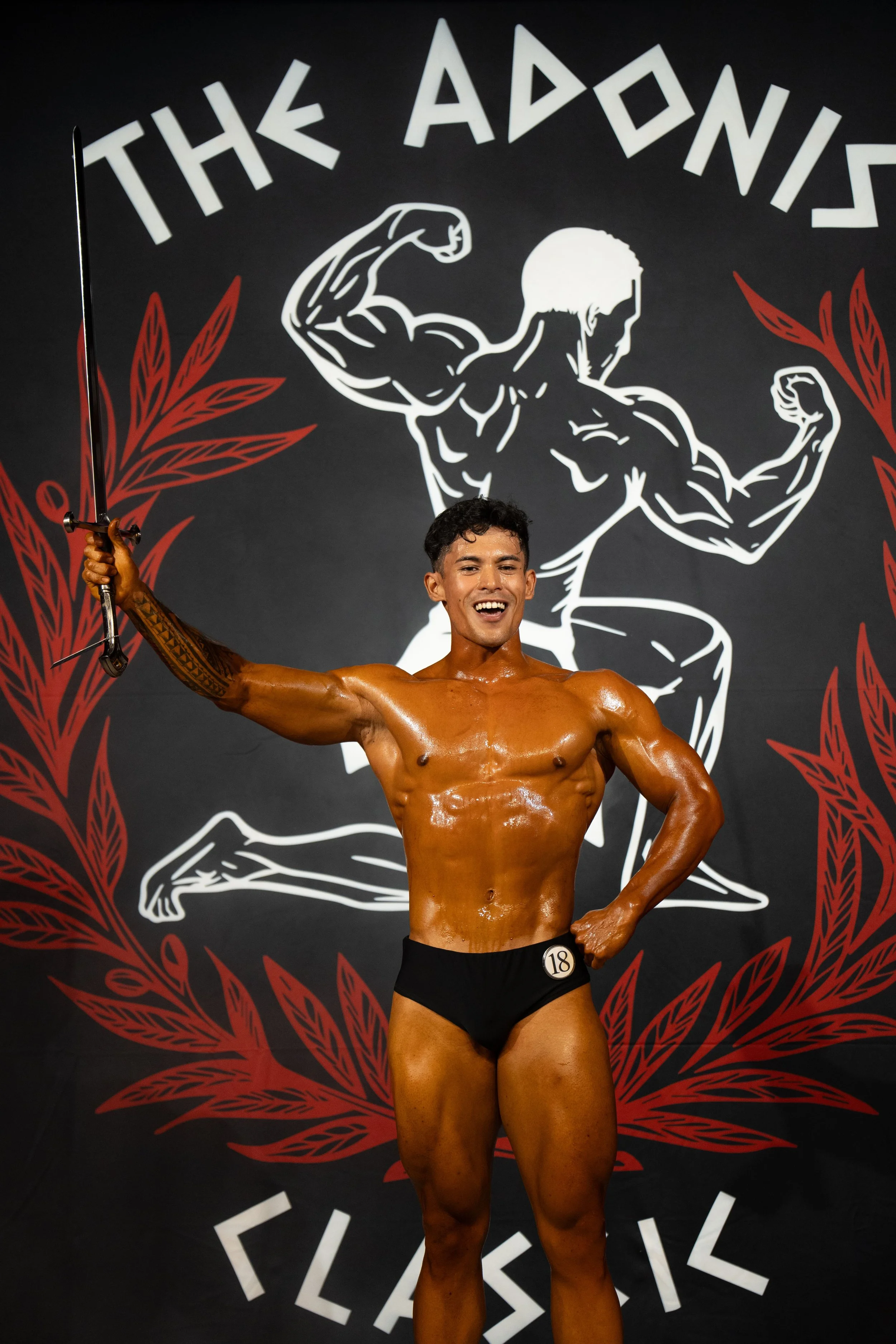A male bodybuilder with tanned, muscular physique posing on stage during a competition, wearing black posing trunks with a number 18 badge, holding a sword in one hand, smiling, standing in front of a backdrop with a muscular figure and text that rea