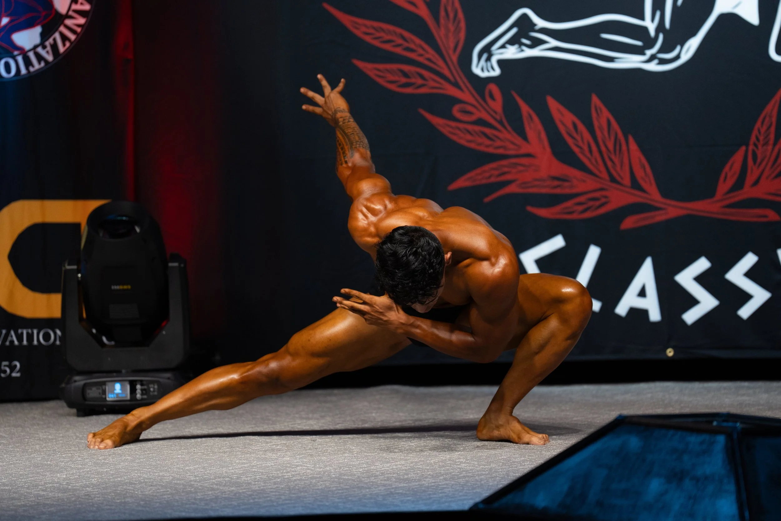 A muscular man with tan skin performing a yoga pose on stage, balancing on one hand with one leg extended and the other bent. He is shirtless, with black curly hair, and standing in front of a black backdrop with red leaves and a sketch of a person i