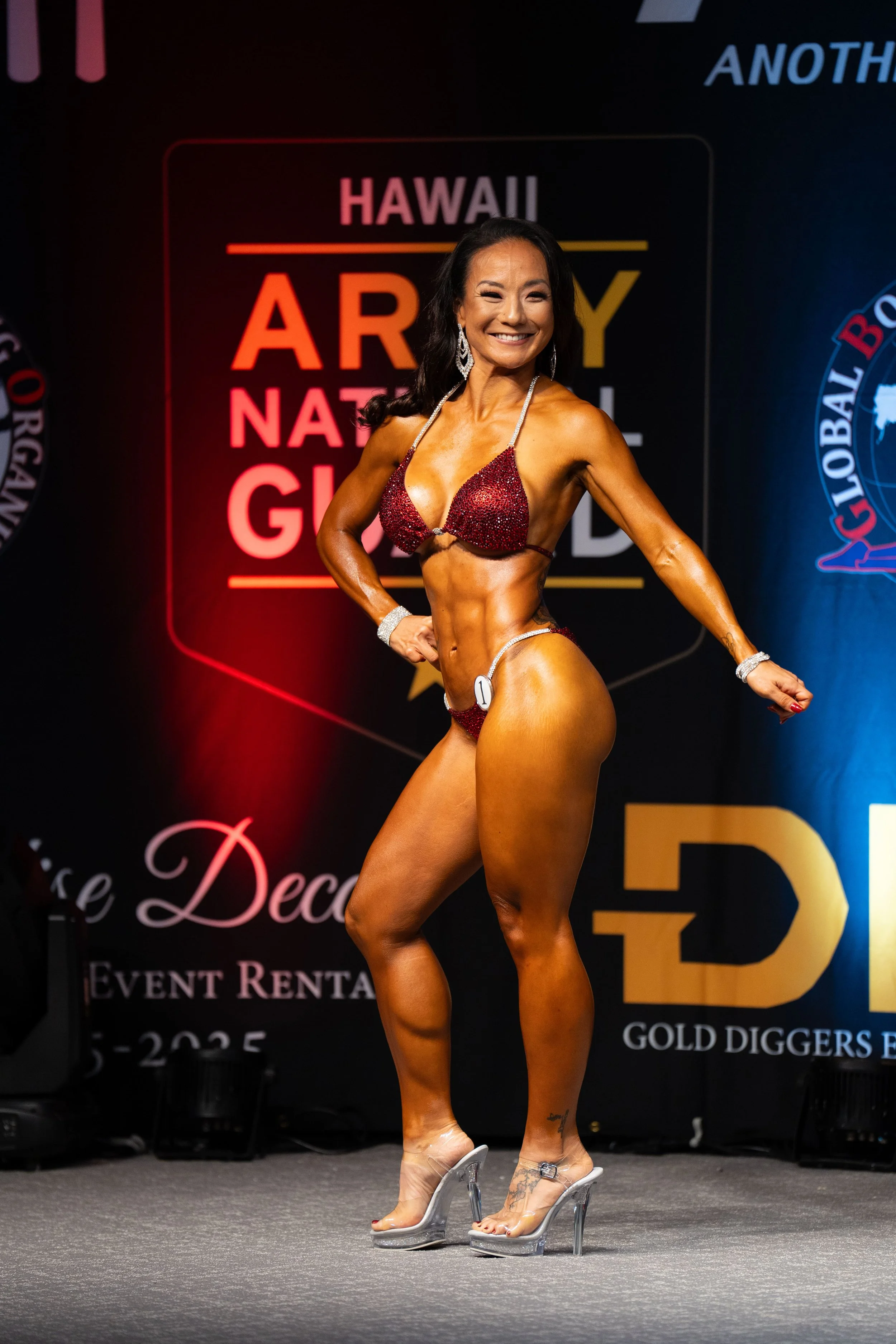 A female bodybuilder on stage in a red bikini, smiling, showcasing her muscular physique at a bodybuilding competition.