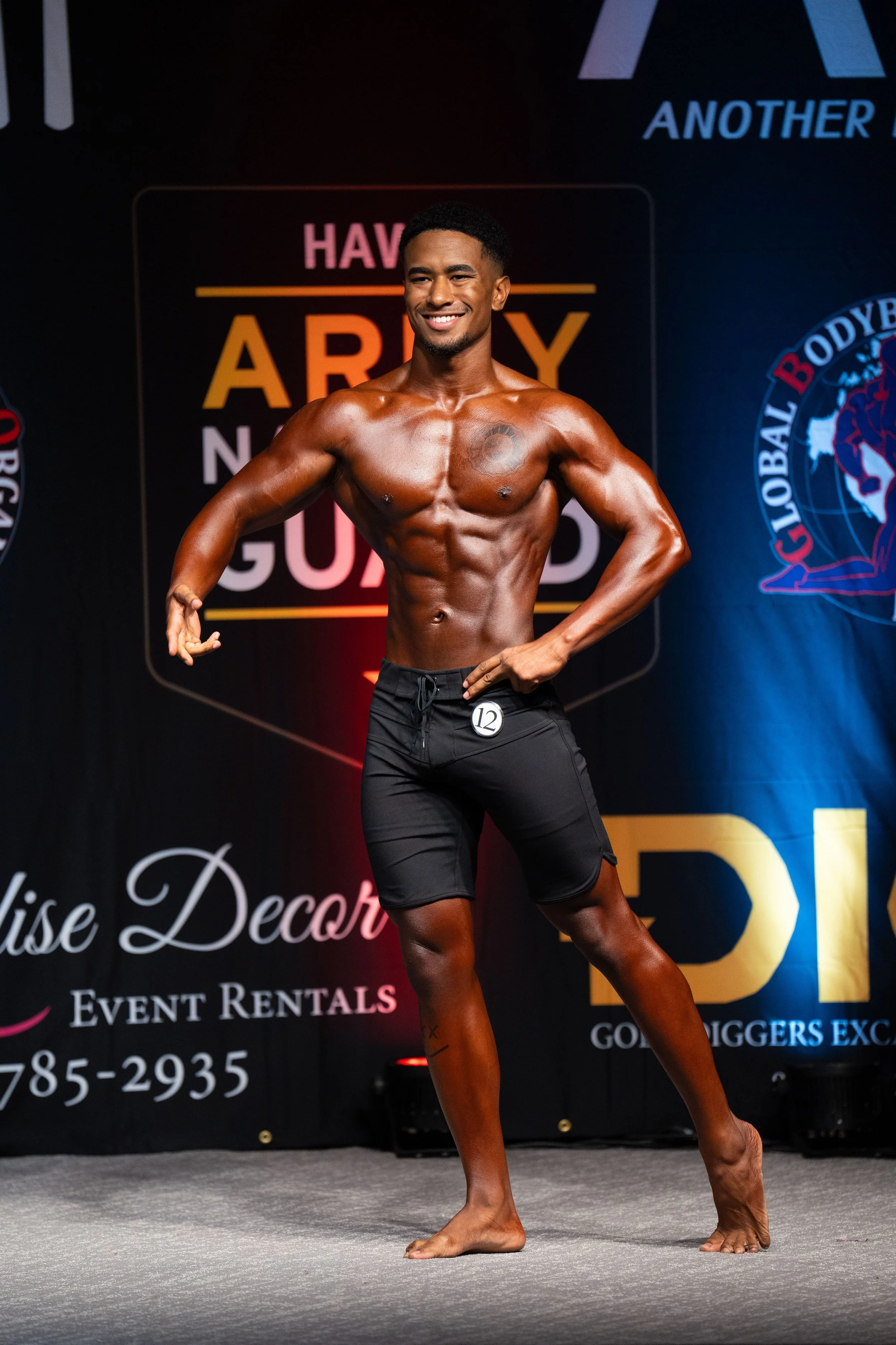 A male bodybuilder on stage at a bodybuilding competition, posing with one arm flexed and smiling. He is wearing black shorts and has a contestant number 12 attached to his shorts. The background features a black curtain with colorful logos and text.