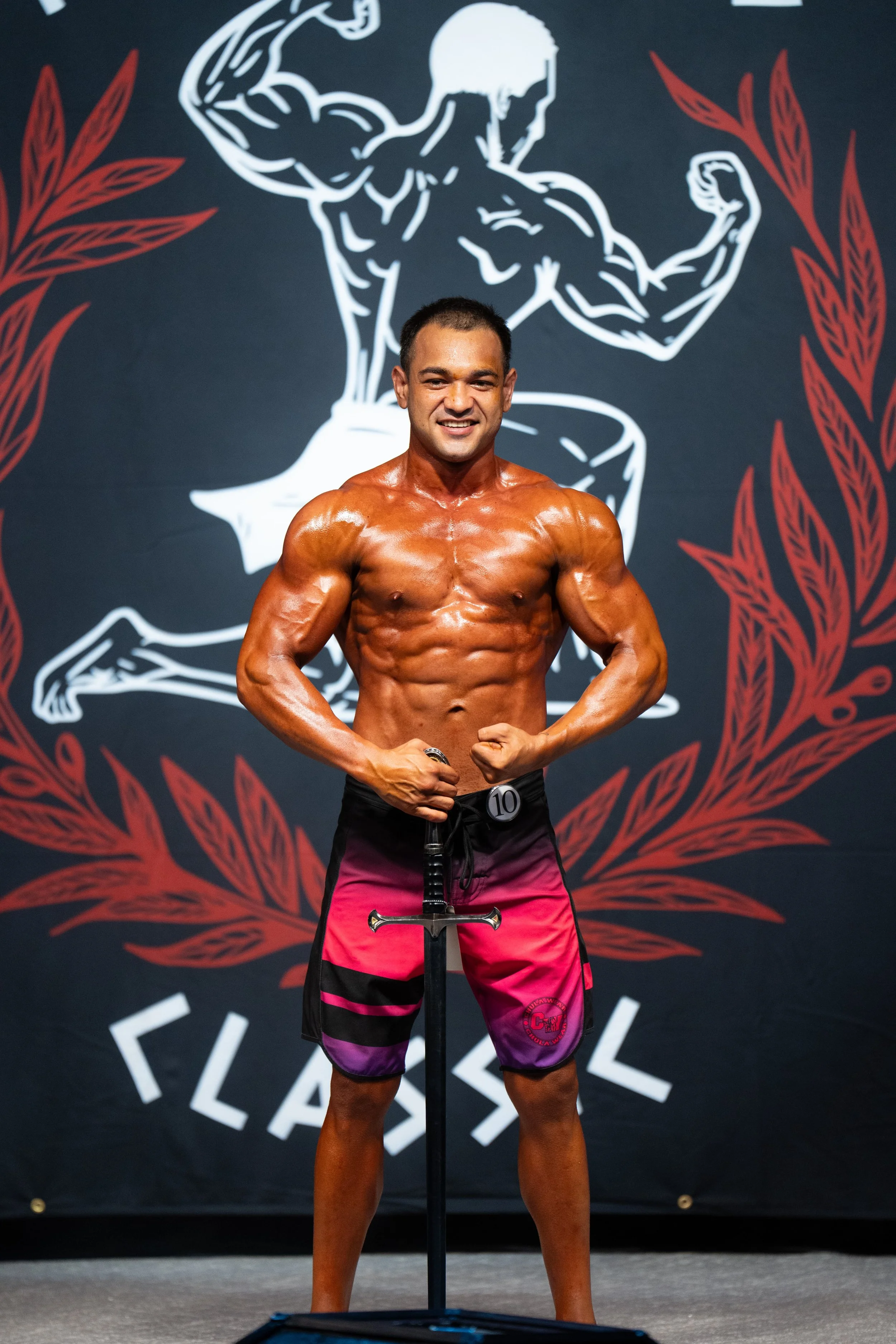 A muscular man standing on stage in a bodybuilding competition, holding a sword vertically in front of him. He is shirtless, wearing pink and black shorts, and smiling. Behind him is a large black backdrop with a white outline of a muscular figure an
