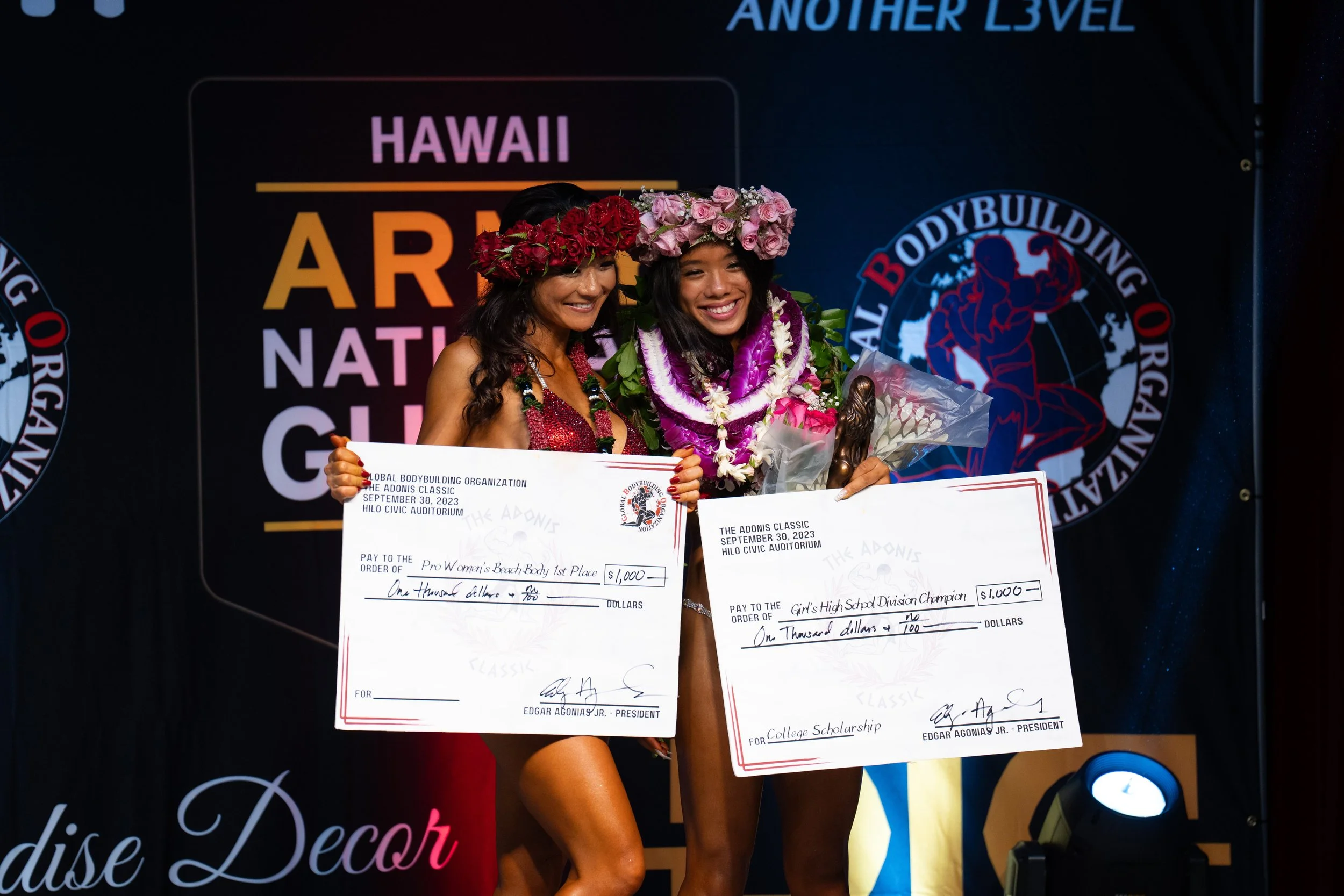Two young women dressed in fitness attire, wearing flower crowns and leis, holding oversized checks and smiling on stage at a bodybuilding competition awards ceremony.