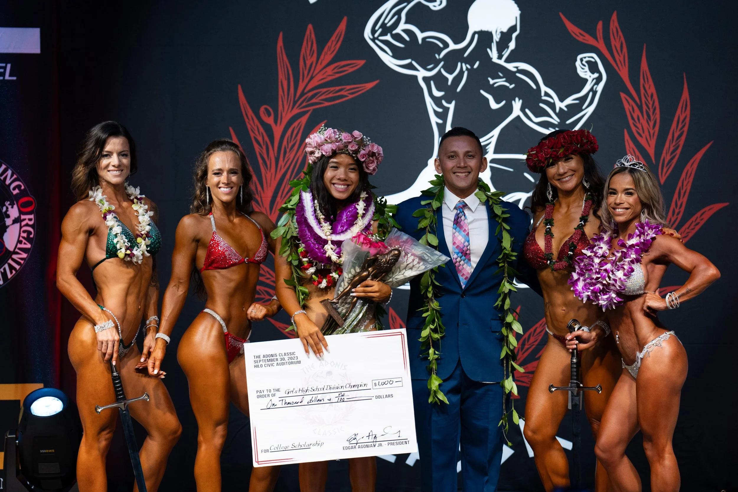 Bodybuilding competition winners on stage, women in bikinis with leis and flowers, one holding a large check and bouquet, man in suit with lei, large muscular logo in background.