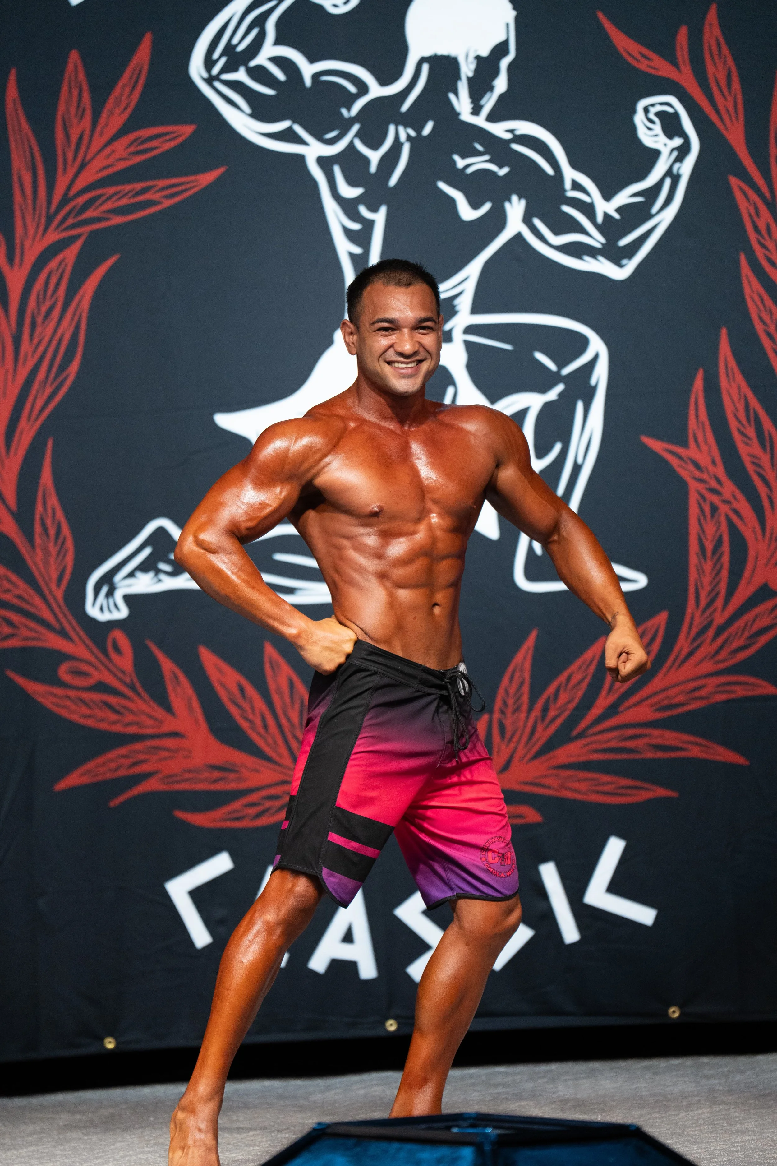 A muscular man in colorful shorts posing on stage at a bodybuilding competition with a backdrop featuring a large graphic of a flexing bodybuilder and red decorative elements.