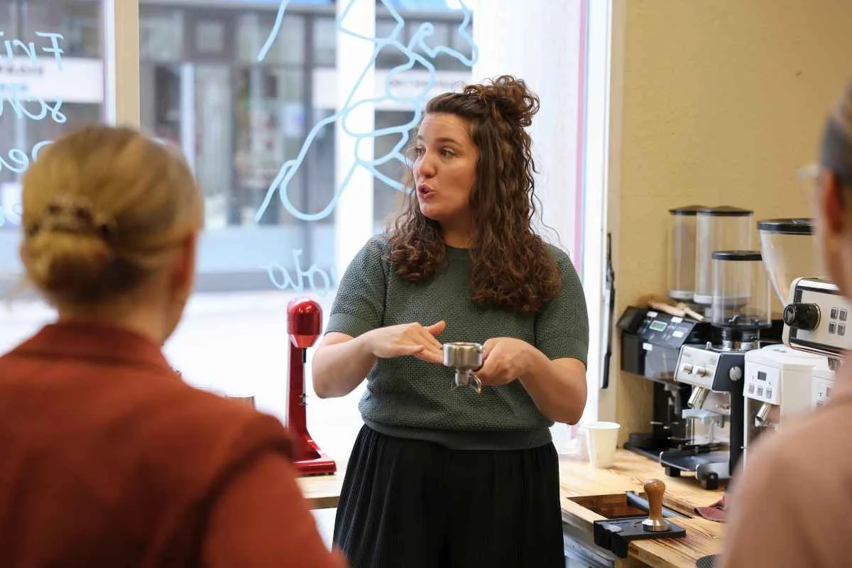 A woman with curly brown hair, wearing a green top, is speaking to two women inside a coffee shop. The woman is gesturing with her hands and holding a coffee portafilter. The coffee shop has coffee machines and grinders on the counter behind her, and a window with writing in the background.