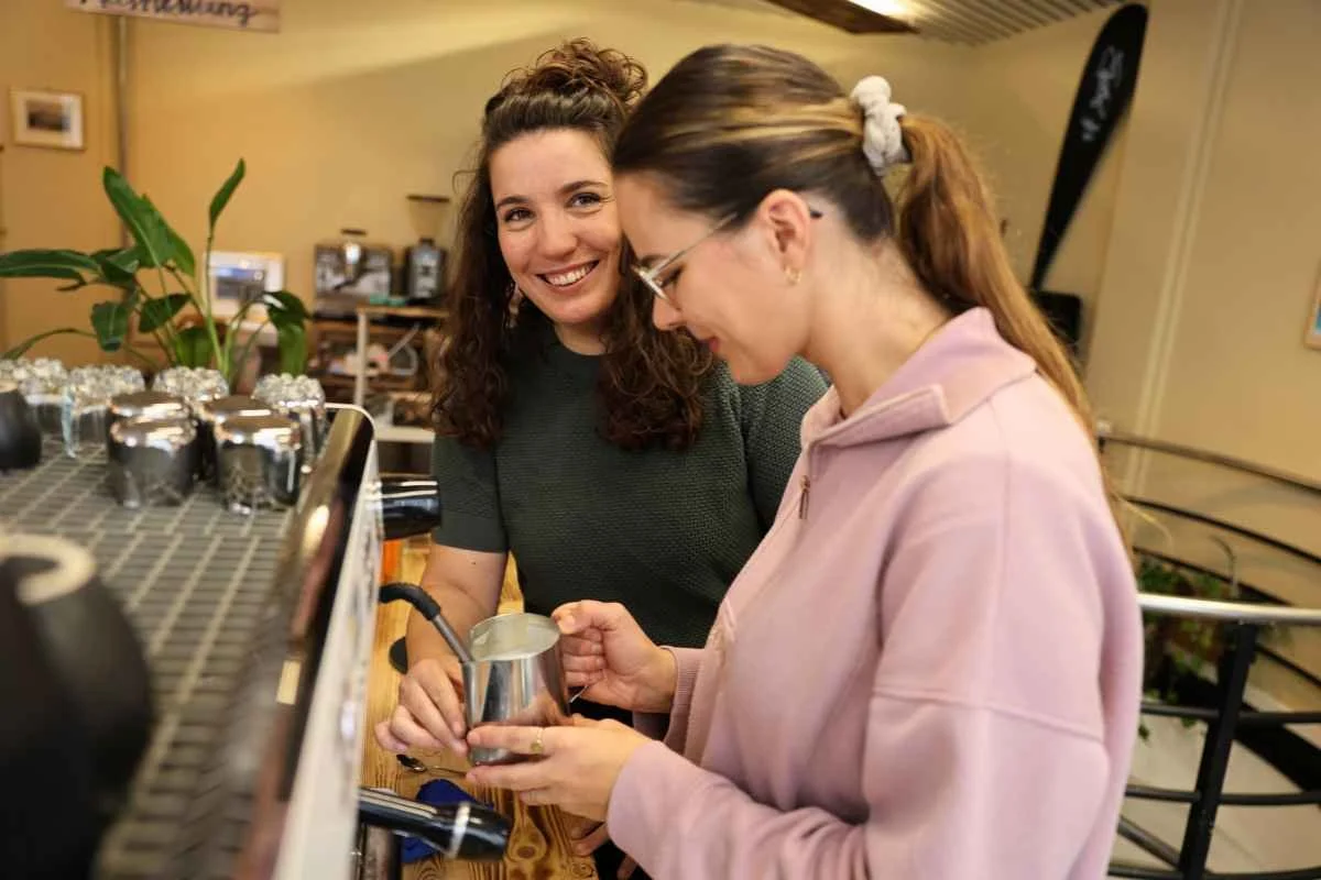 A Barista teacher teaches a woman how to steam milk for latte art in a cozy cafe, smiling and engaged in making coffee.