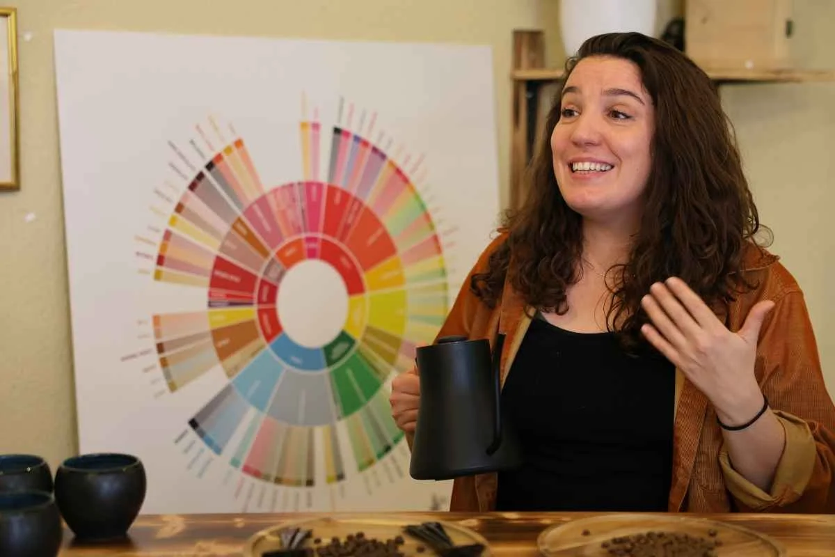A woman with curly brown hair, wearing a green top, is speaking to two women inside a coffee shop. The woman is gesturing with her hands and holding a coffee portafilter. The coffee shop has coffee machines and grinders on the counter behind her, and a window with writing in the background.