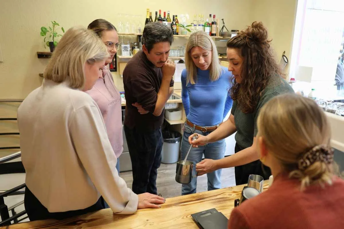 A Barista teacher teaches a woman how to steam milk for latte art in a cozy cafe, smiling and engaged in making coffee.