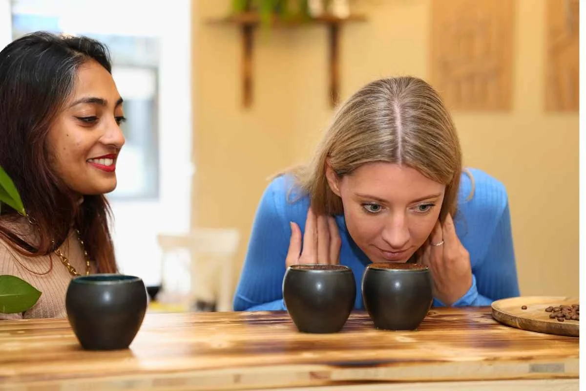 Two women tasting coffee from black cups on a wooden table in a cozy room.