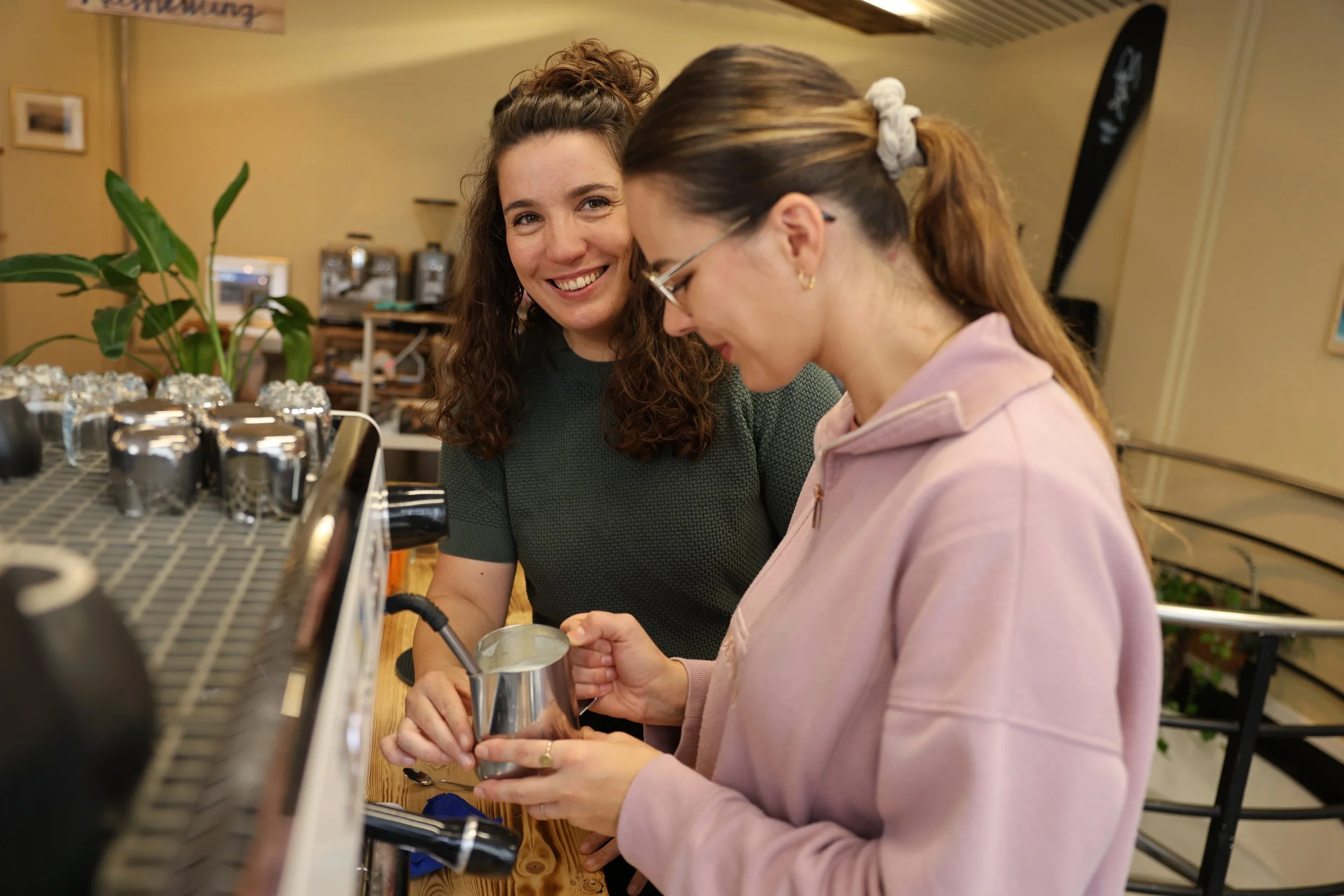 A Barista teacher teaches a woman how to steam milk for latte art in a cozy cafe, smiling and engaged in making coffee.