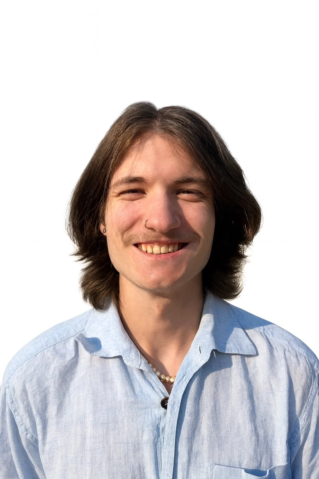 Portrait of a smiling young person with shoulder-length brown hair, wearing a light blue button-up shirt, a pearl necklace, and a nose ring, against a plain white background.