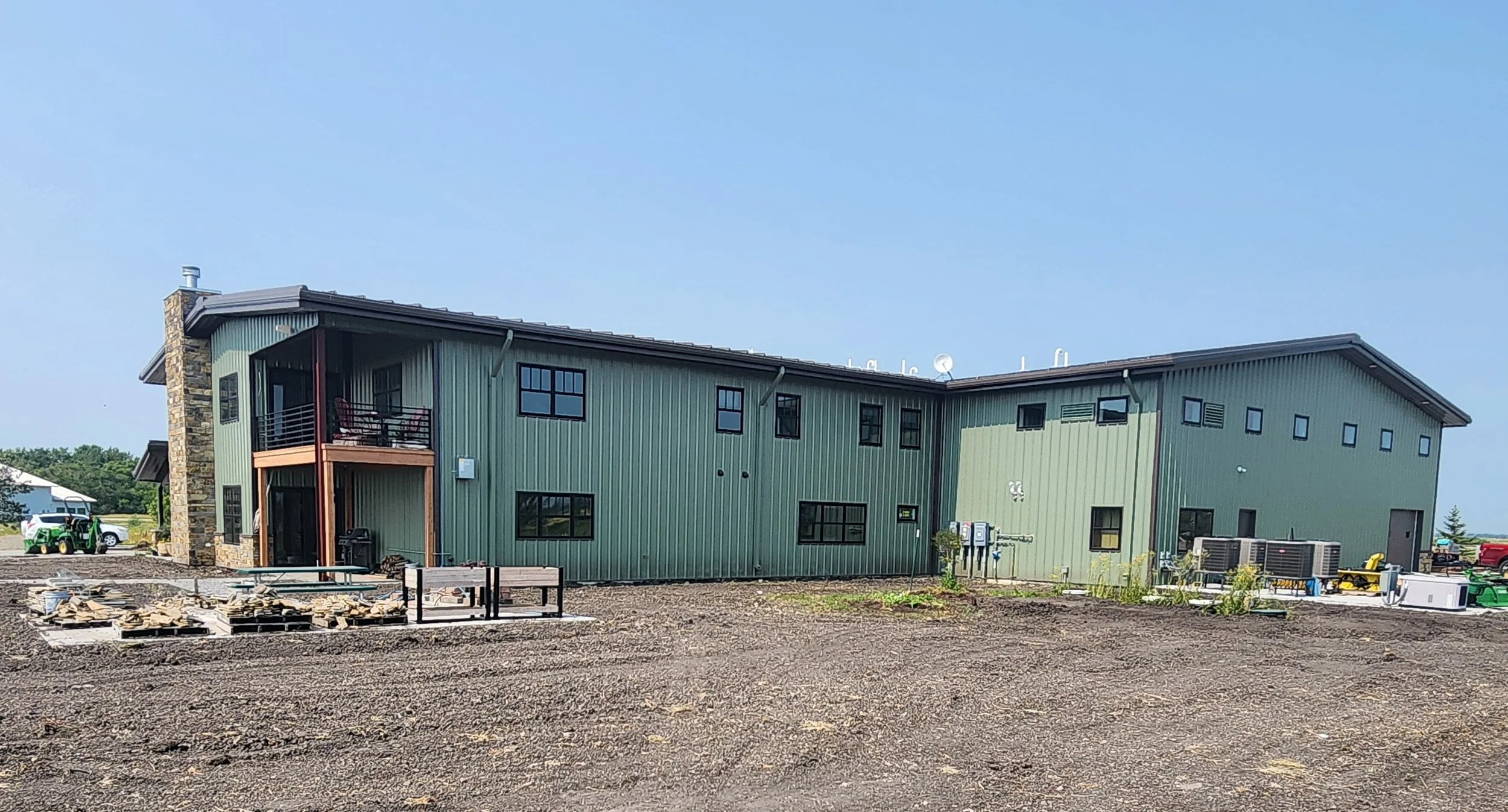 Green modern residential building with stone chimney, multiple windows, small balcony with outdoor furniture, situated in a rural setting with construction materials and equipment in the foreground.