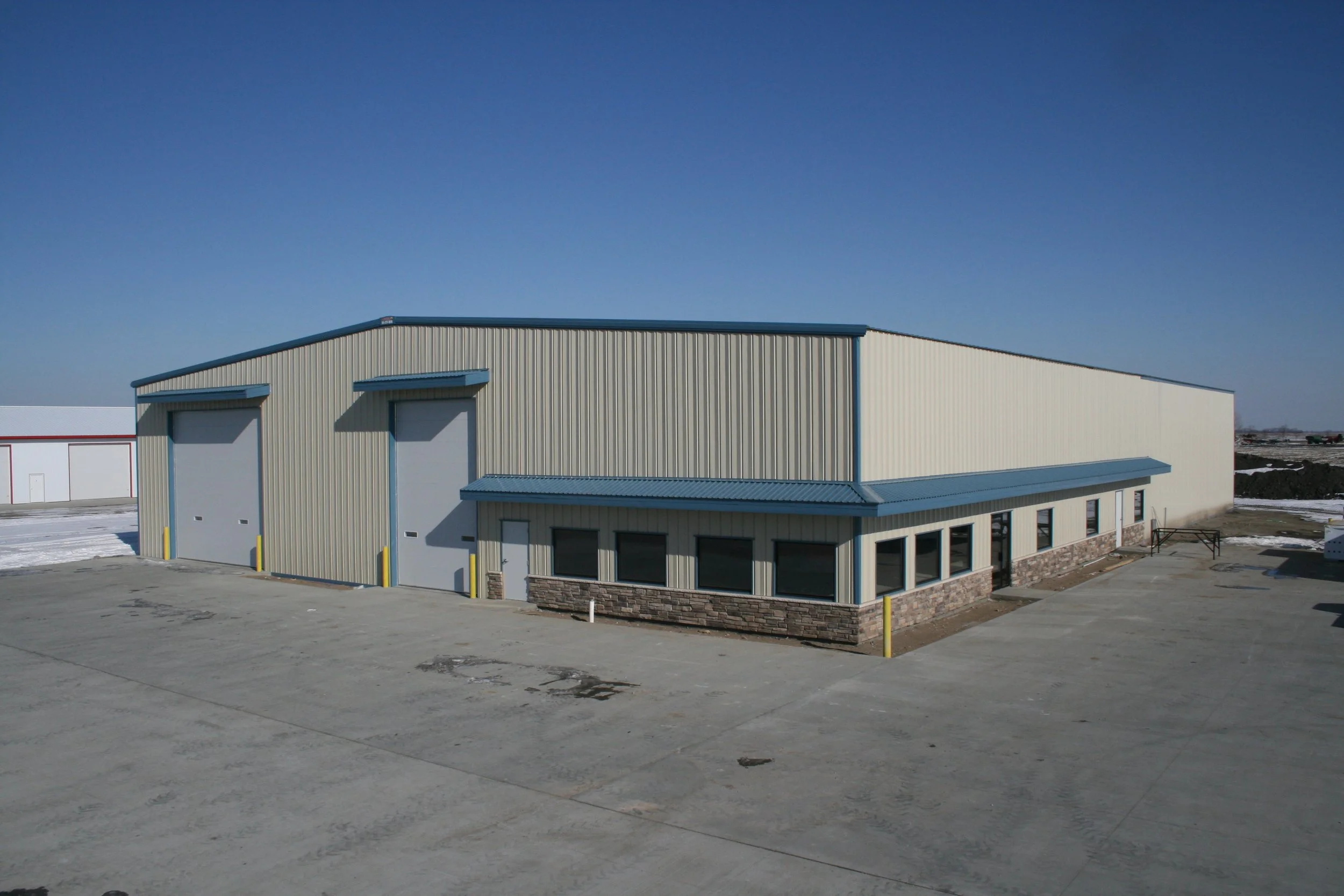 Large industrial building with beige metal siding, blue trim, and two garage doors, located on a concrete lot with no vehicles or people around.