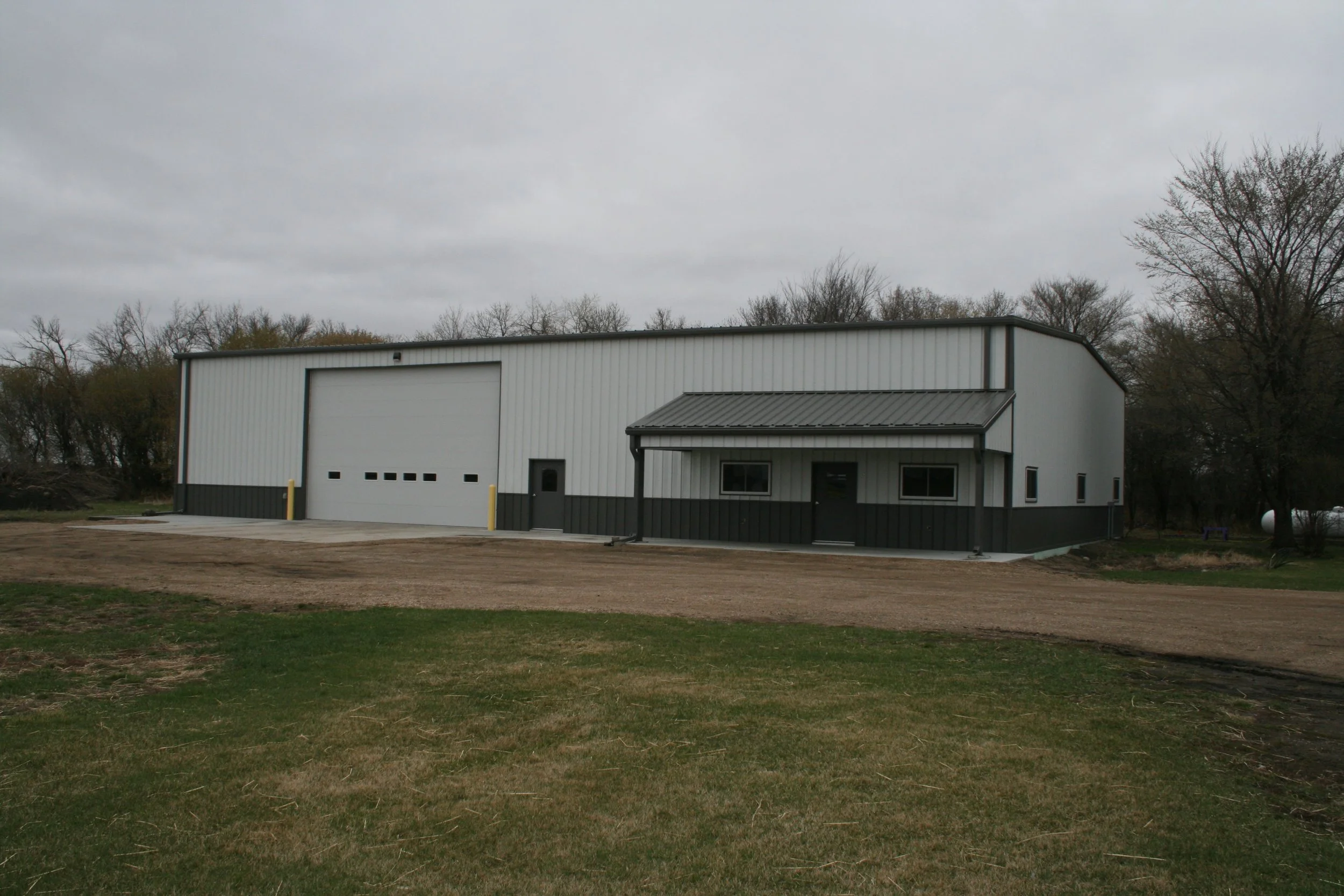 A modern metal building with a large garage door, smaller door, and small windows, situated on a patch of dirt and grass, with trees and overcast sky in the background.