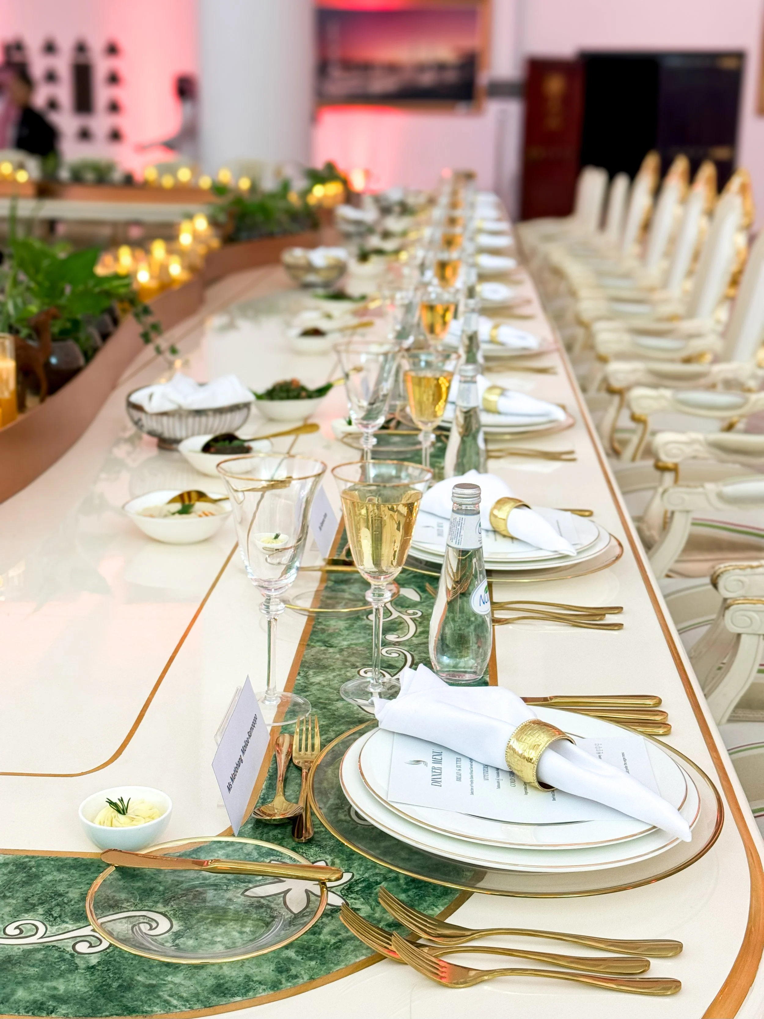 Elegant banquet table set with gold cutlery, white plates with gold rims, white napkins with gold rings, and glasses of white wine. Small bowls of condiments and greenery decor line the center of the table, with chairs arranged along the sides in a well-lit room.