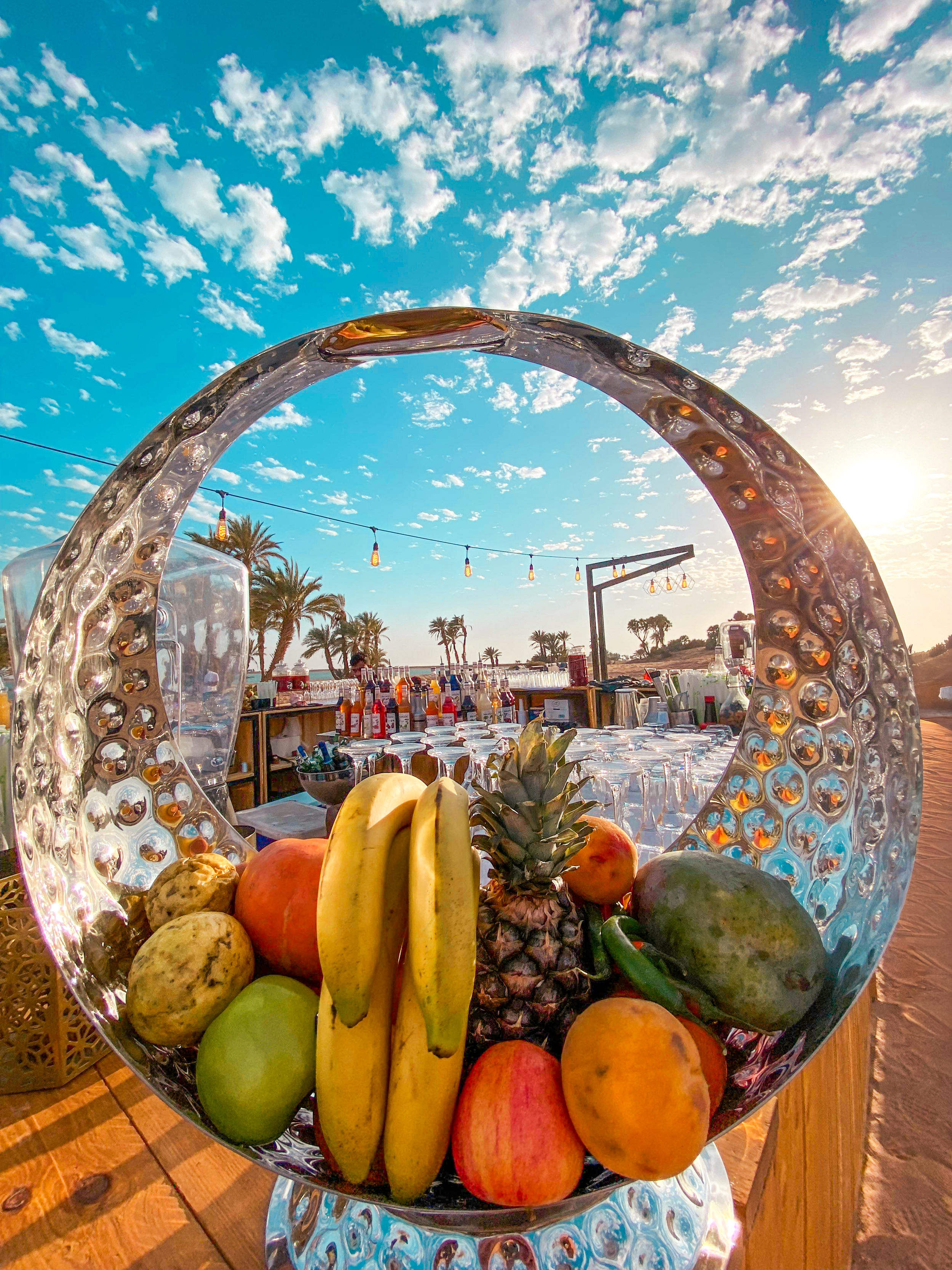 Fruits in a clear glass bowl, with bananas, pineapple, apples, watermelon, and oranges, set on a wooden table outdoors at sunset with a sky filled with clouds and palm trees in the background.