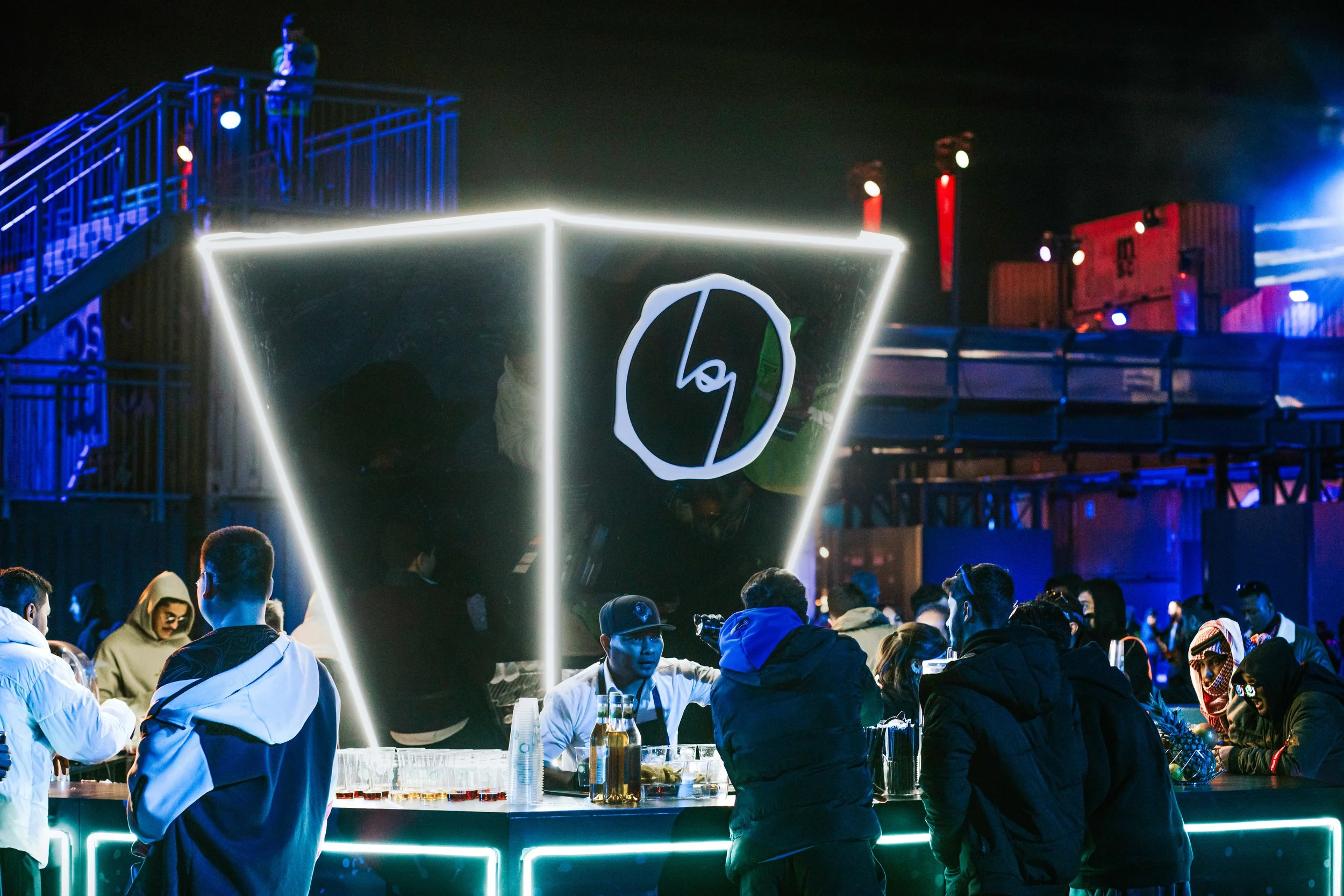 Nighttime scene at a festival with people gathered around a glowing bar with a cube-shaped reflective surface, illuminated by neon lights, with stacked shipping containers and stage lights in the background.
