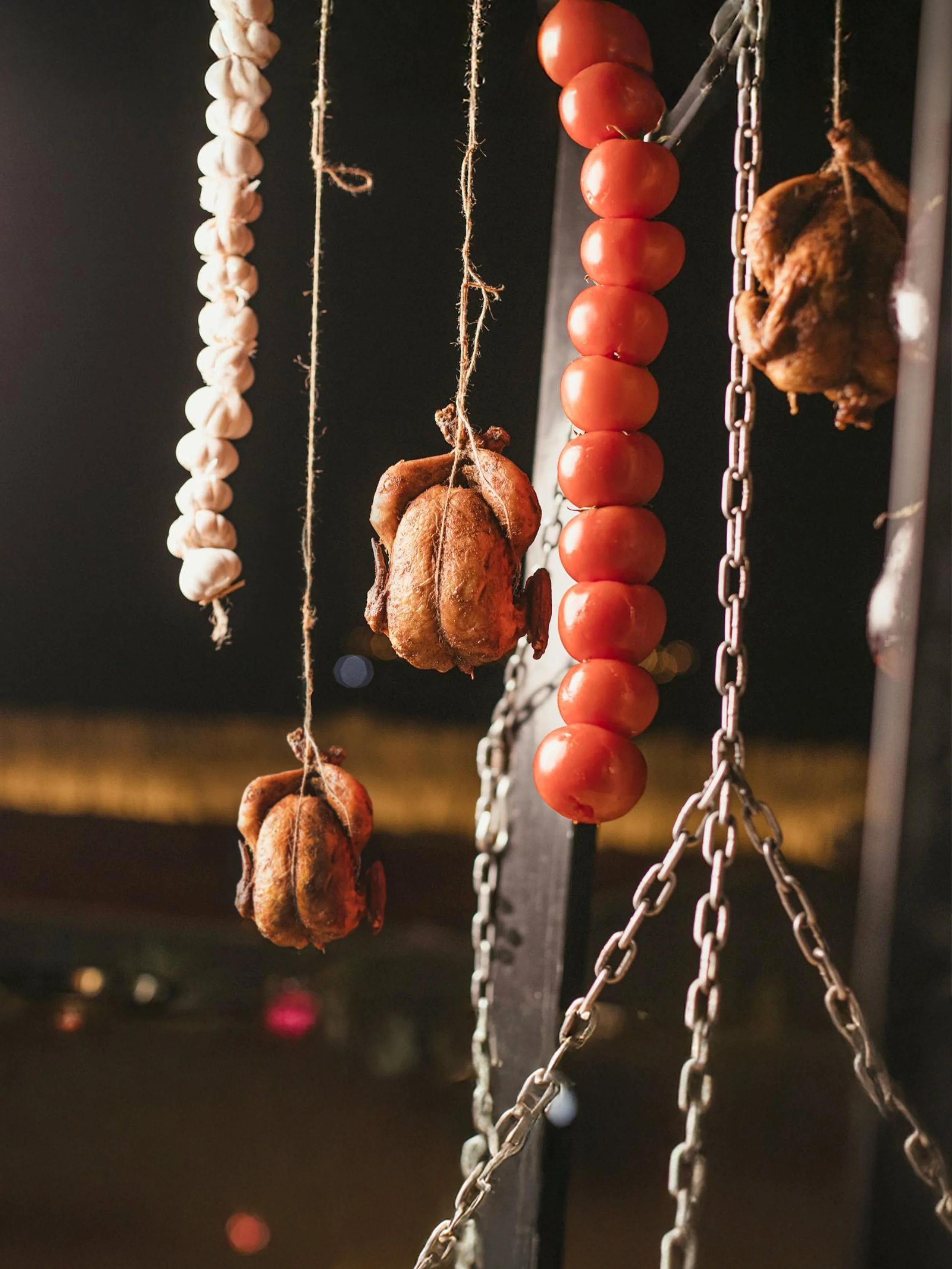 Hanging skewers of garlic, cherry tomatoes, and roasted chicken pieces on chains.