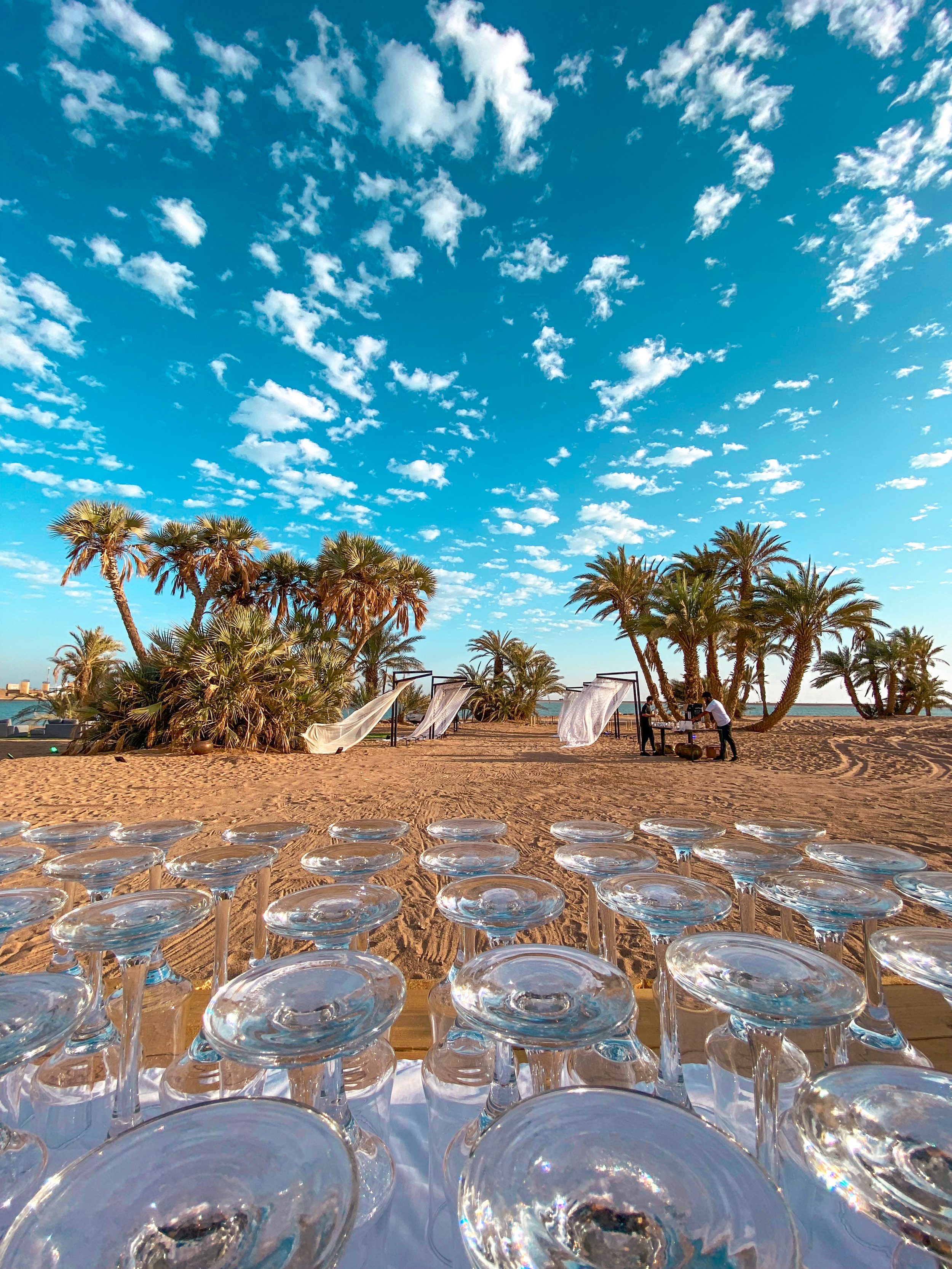 A beach setup with several empty glasses on a table in the foreground, sand, palm trees, and cabanas in the background, under a blue sky with scattered clouds.
