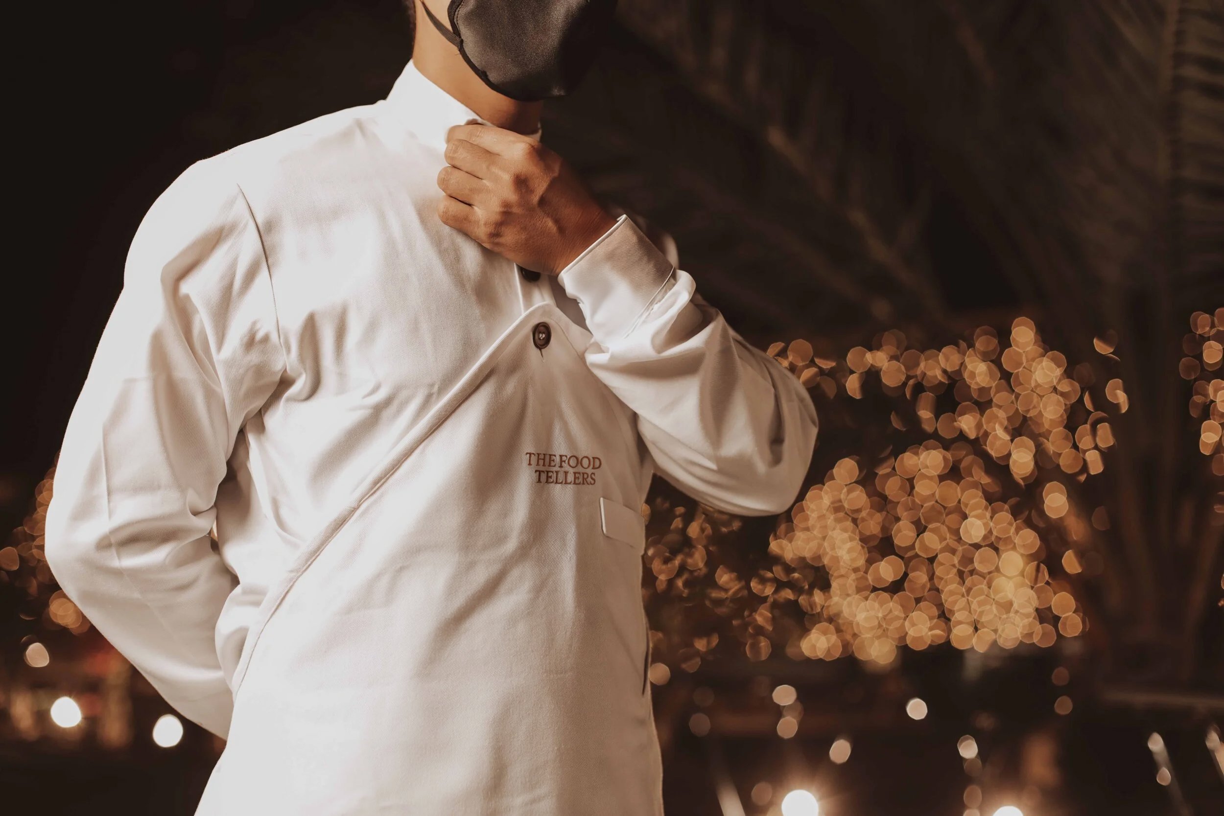 A person wearing a white chef's coat with the text 'THE FOOD TELLERS' on it, adjusting their black face mask, with blurred string lights in the background.