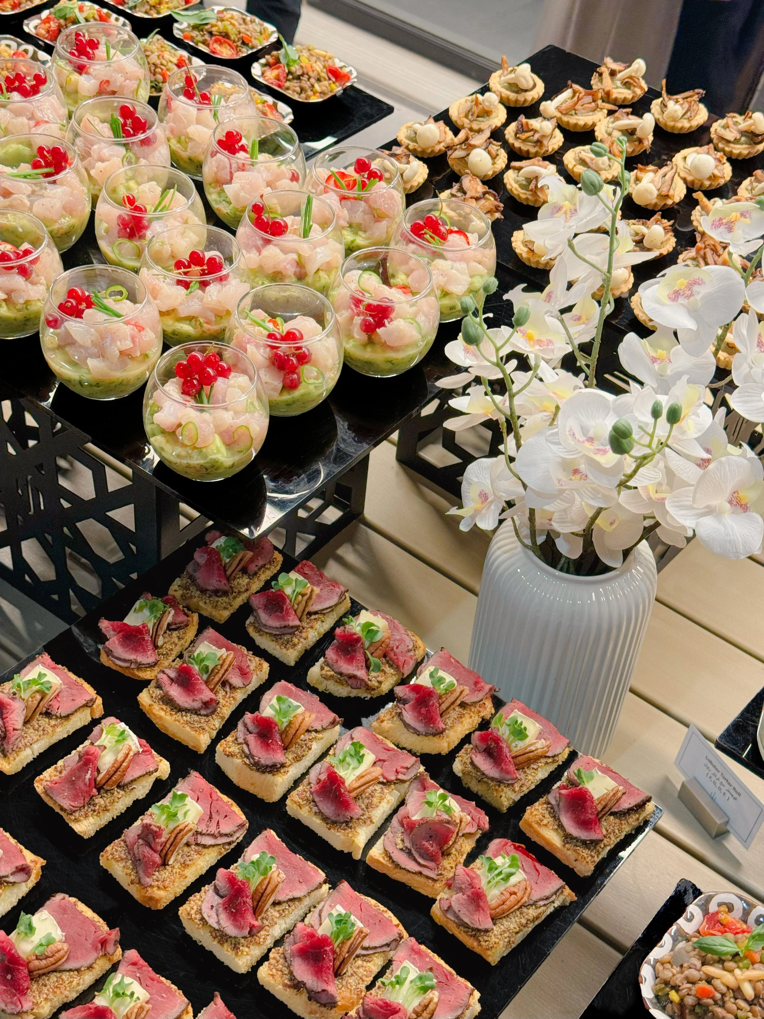 Assorted appetizers and desserts including small cups of seafood with red berries, topped with green garnish; small tartlets with cream and toppings; small canapé sandwiches with various toppings; and a white vase with white orchids.