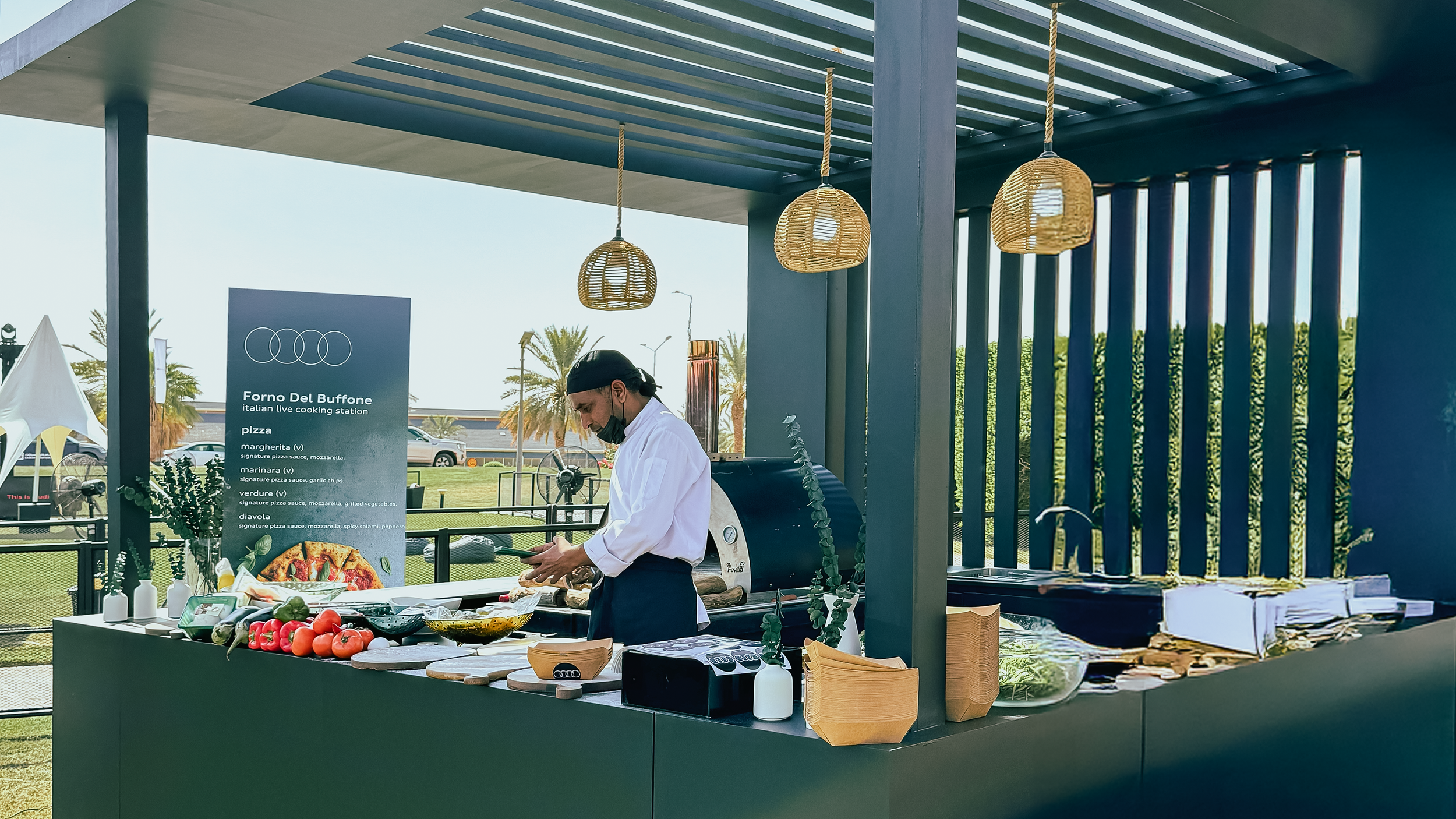 A chef working at an outdoor Italian live cooking station with fresh vegetables, a pizza oven, and a menu sign.