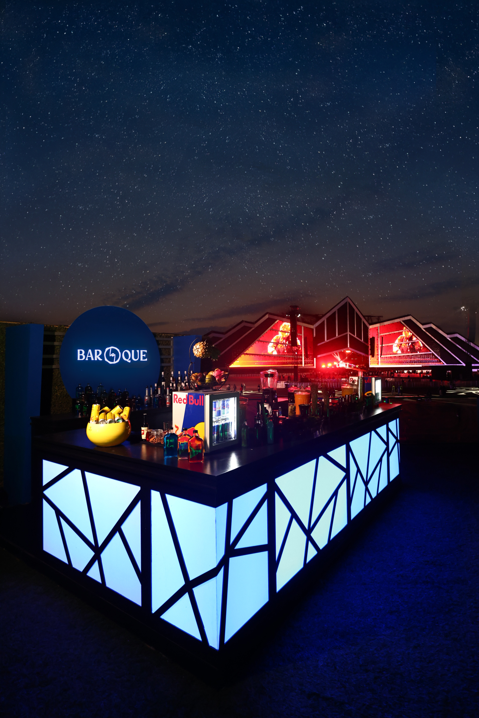 A nighttime view of an outdoor bar with illuminated geometric patterned panels, set against a dark sky filled with stars, and a modern building with red lighting in the background.