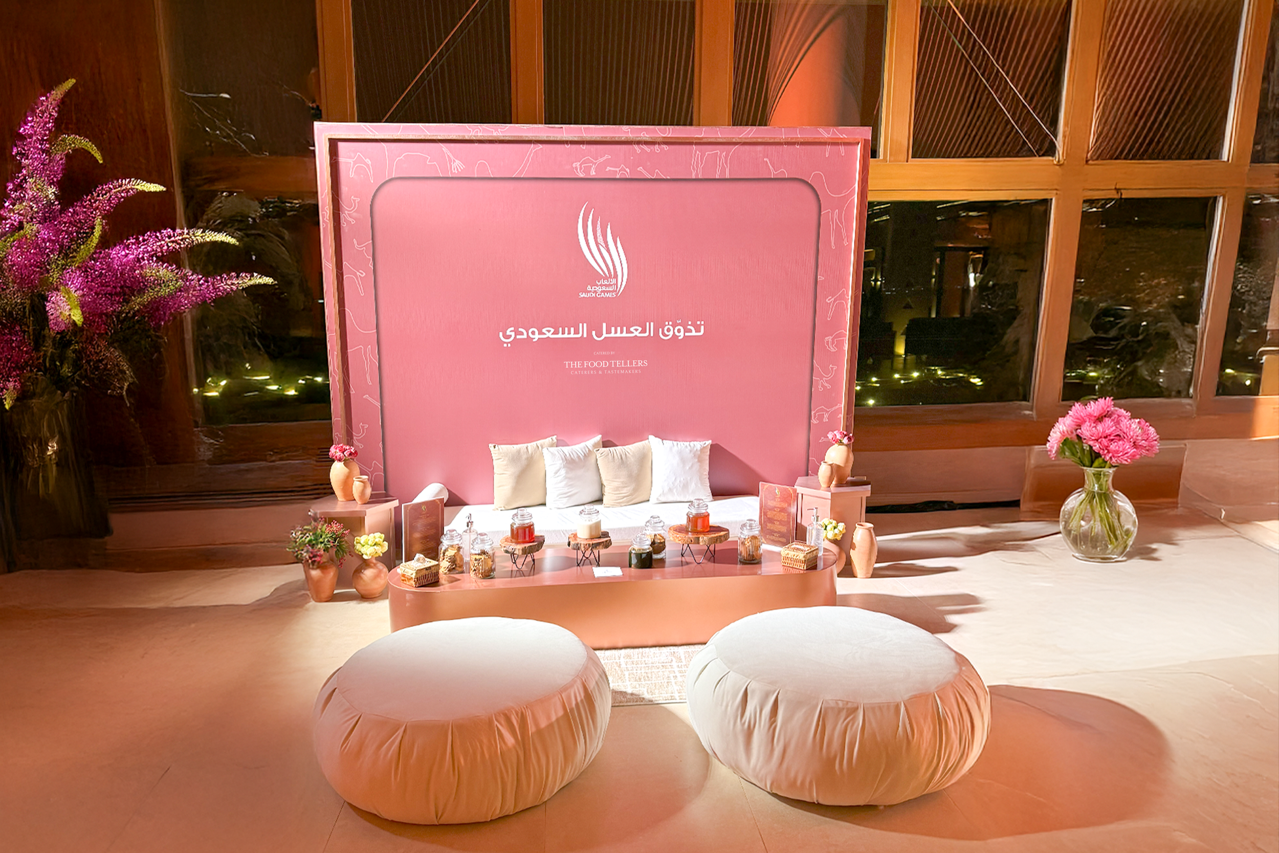 Elegant pink display with cushions and decorative jars, set against a pink backdrop with Arabic writing, flowers, and vases, near large windows at night.