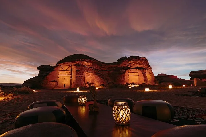 Outdoor dining setup during sunset with candlelit tables, desert landscape, and rock formations in the background.