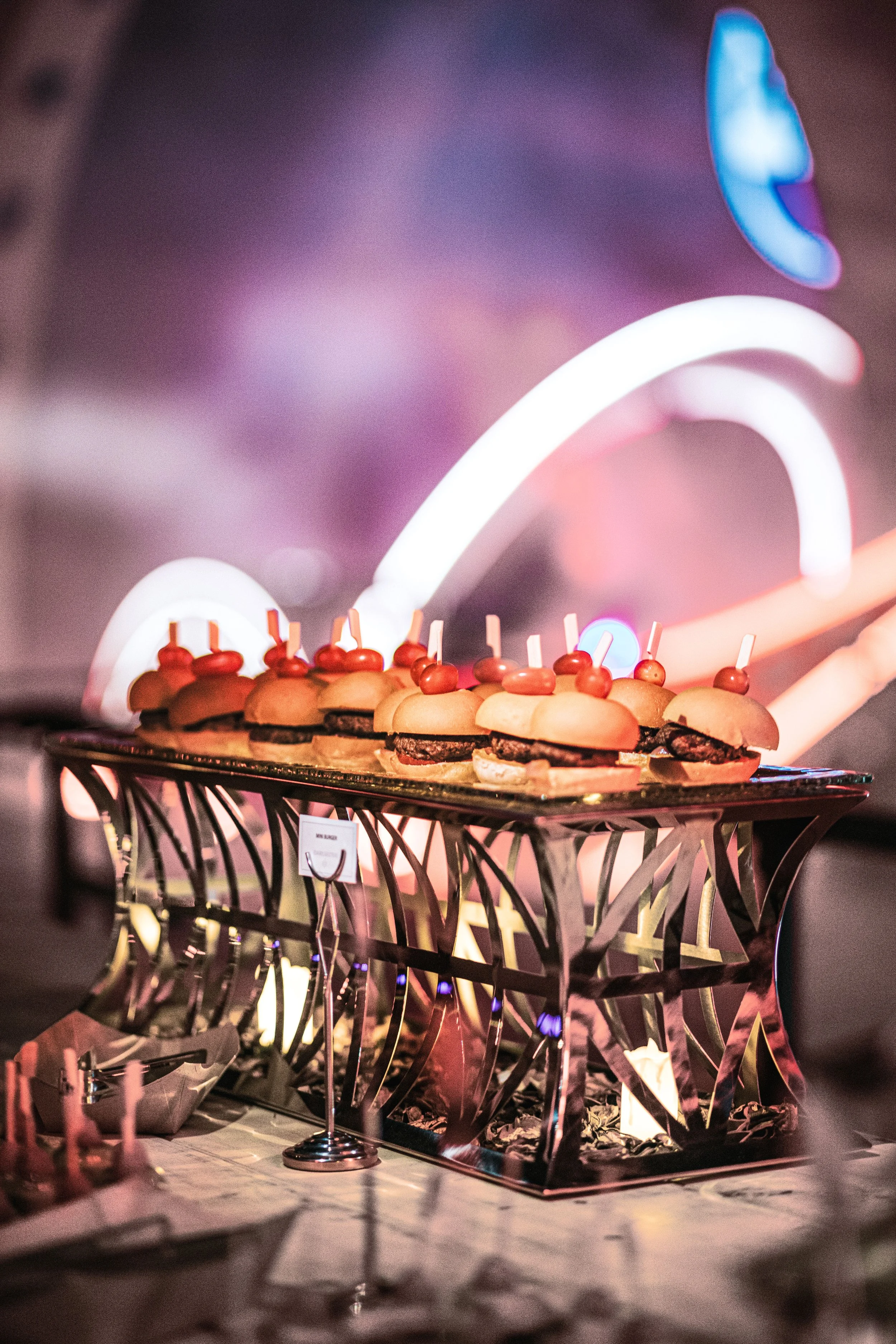 Tray of mini sliders with cherry tomatoes on skewers at a buffet with neon lights in the background.